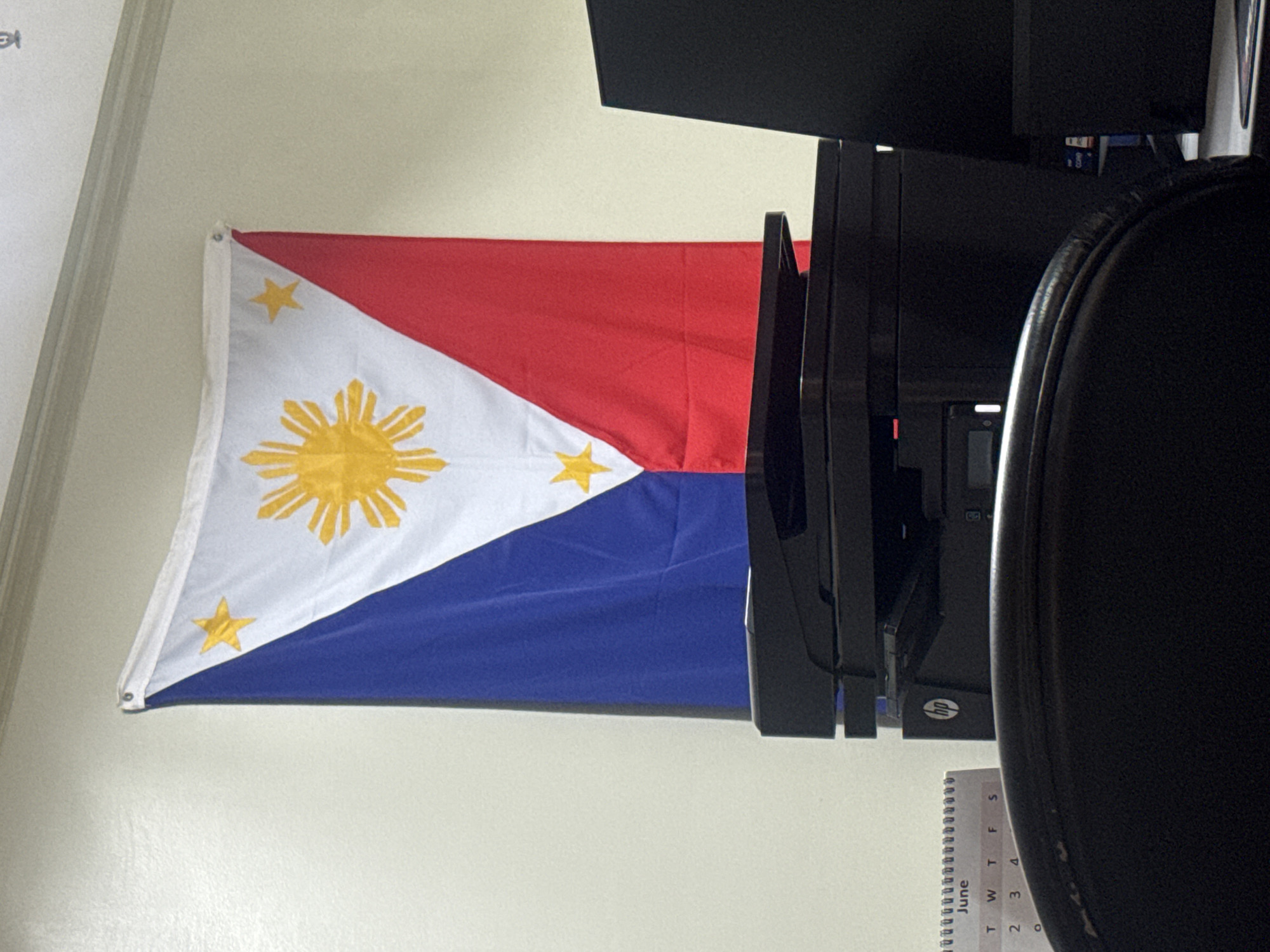 The image shows a Philippine flag hanging on a white wall. The flag is predominantly white with a blue and red stripe on the left and right sides respectively. A yellow sun with radiating rays is at the center of the white triangle, and three yellow stars are at each corner of the triangle. In front of the flag, on a desk, are a black printer, a black computer monitor, and a black office chair. A desk calendar with "June" visible at the top is also on the desk. The lighting suggests an indoor setting, possibly an office. The image does not provide any specific details about the city of Davao City, Philippines.
