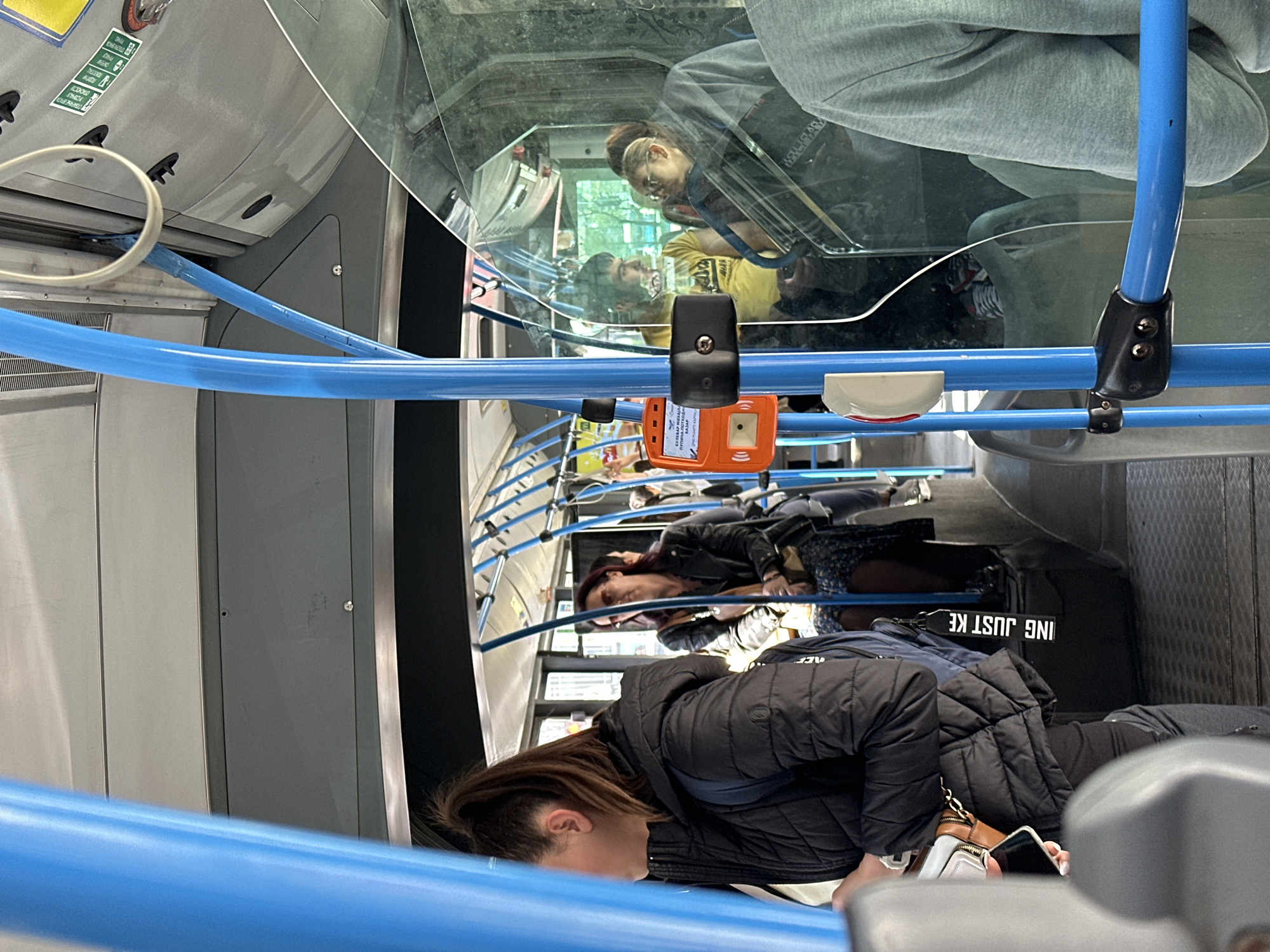 The image depicts the interior of a bus in Novi Sad, Serbia. Several passengers are present, with at least three visible and more partially seen. The first passenger, closest to the camera, is a woman wearing a black puffer jacket with her head tilted downwards and a phone in her hand. Behind her, another woman in a black jacket and floral dress is seated. A black bag with the text "ING JUST KE" is visible next to her. Further into the bus, other passengers are seated, and a reflection of a man in a yellow shirt can be seen in the window. Blue handrails run along the ceiling of the bus. An orange electronic ticket reader is mounted on one of the handrails. The bus interior appears modern and well-maintained.