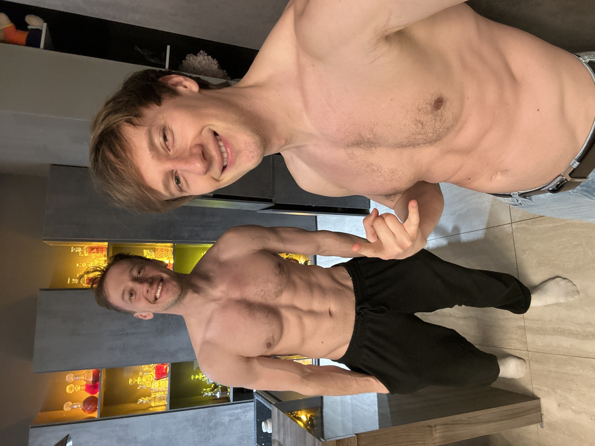 Two shirtless men are posing indoors. The man on the left wears jeans and a belt. The man on the right wears black pants and white socks. Behind them is a kitchen area with glass bottles visible inside cabinets. The location is the city of Kursk, Russia.