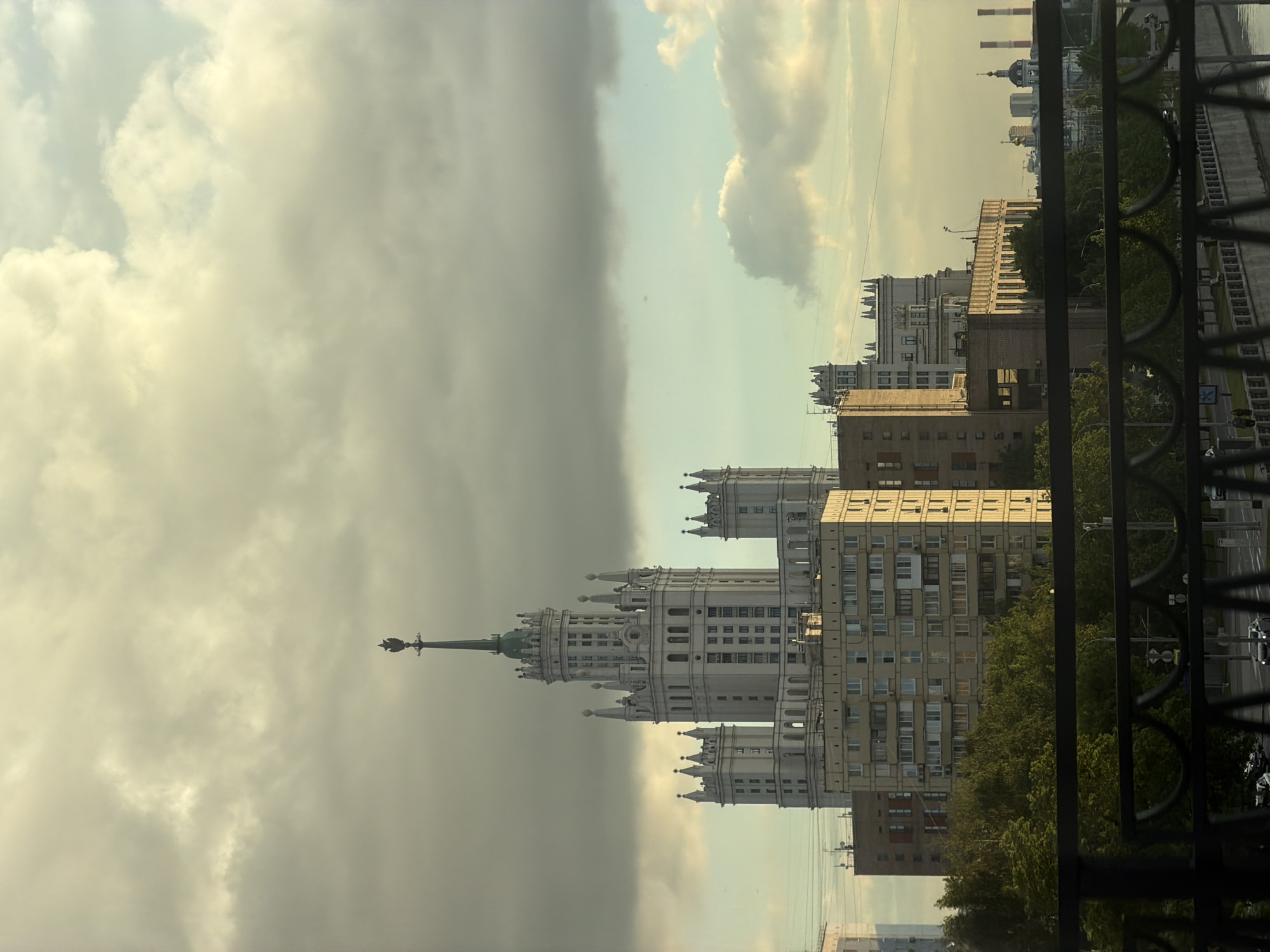The image shows a view of the Kotelnicheskaya Embankment Building in Moscow, Russia, from an elevated perspective. The Stalinist skyscraper is prominently featured in the center. A storm cloud is positioned to the left, while the right is brighter. Other buildings are visible to the right, along with trees and a bridge with a railing in the foreground.