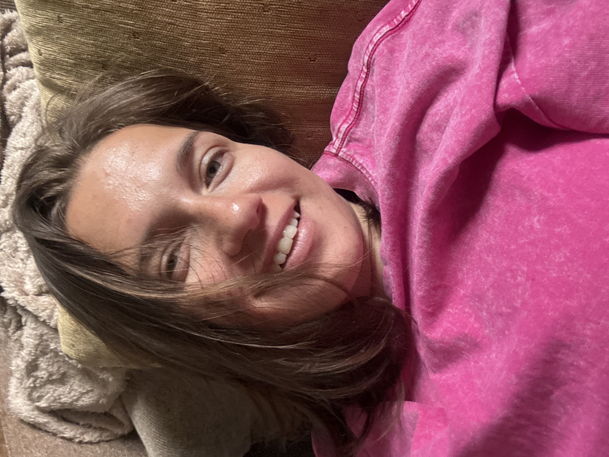 A young woman with brown hair and fair skin is lying down, smiling at the camera. She is wearing a bright pink sweatshirt. She appears to be resting on a sofa or similar upholstered surface. The background shows textures suggesting cushions or blankets. The location is not possible to determine with the given information.