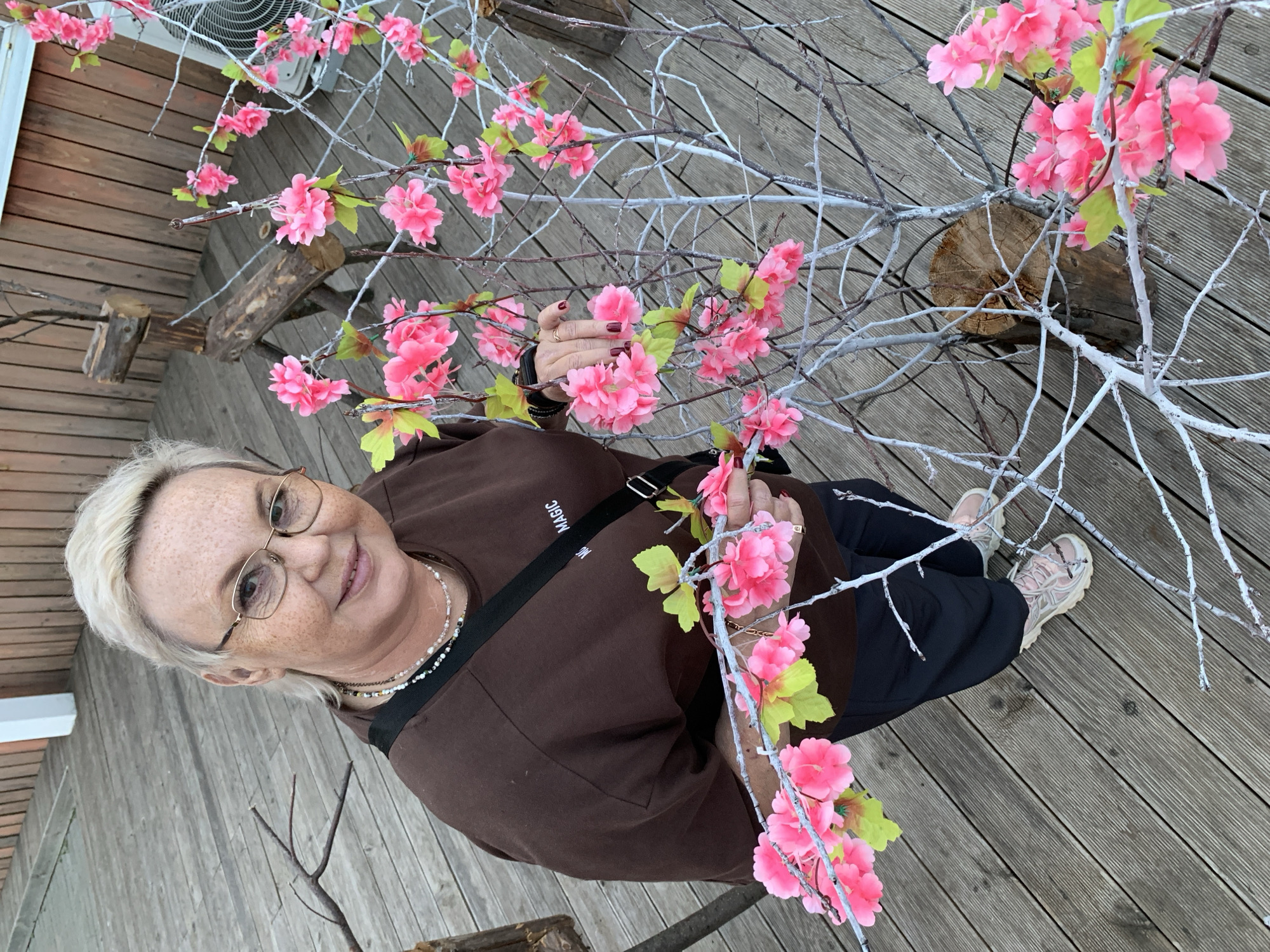 A woman with short blonde hair and glasses, wearing a brown sweatshirt, a beaded necklace, and black pants, poses for a photo outdoors. She is standing on a wooden deck surrounded by artificial branches with pink flowers. In the background, there is a white air conditioning unit and a log. She appears to be smiling and holding a flower branch. The image is taken from a high angle, looking down at the woman. There is no visible text in the image. The setting suggests it could be daytime, with a generally pleasant atmosphere.