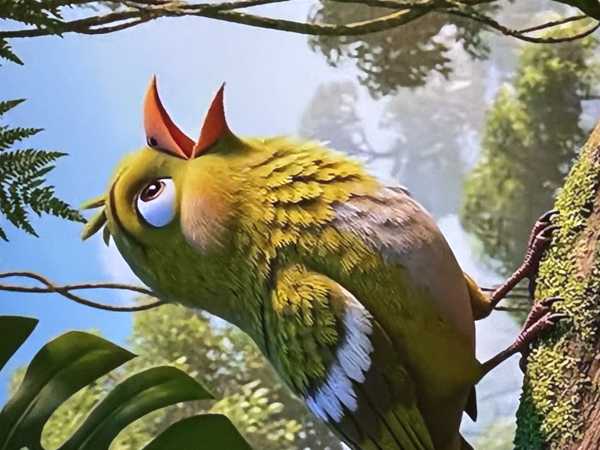 A close-up, eye-level, medium shot captures a stylized, animated green bird perched on a moss-covered branch. The bird is facing right with its beak open, appearing to sing or call. Its large, round blue eye is looking upward and to the right. The bird's body is predominantly green with patches of yellow and a light brown chest. White markings are visible on its wings. The background consists of a blurry, soft-focus depiction of lush green foliage and trees under a bright blue sky, suggesting a jungle or forest environment. Vines hang from the upper right corner. The image is a still from an animation. The provided image does not contain any elements that would allow for its location to be identified in Sabunchu Raion, Azerbaijan.