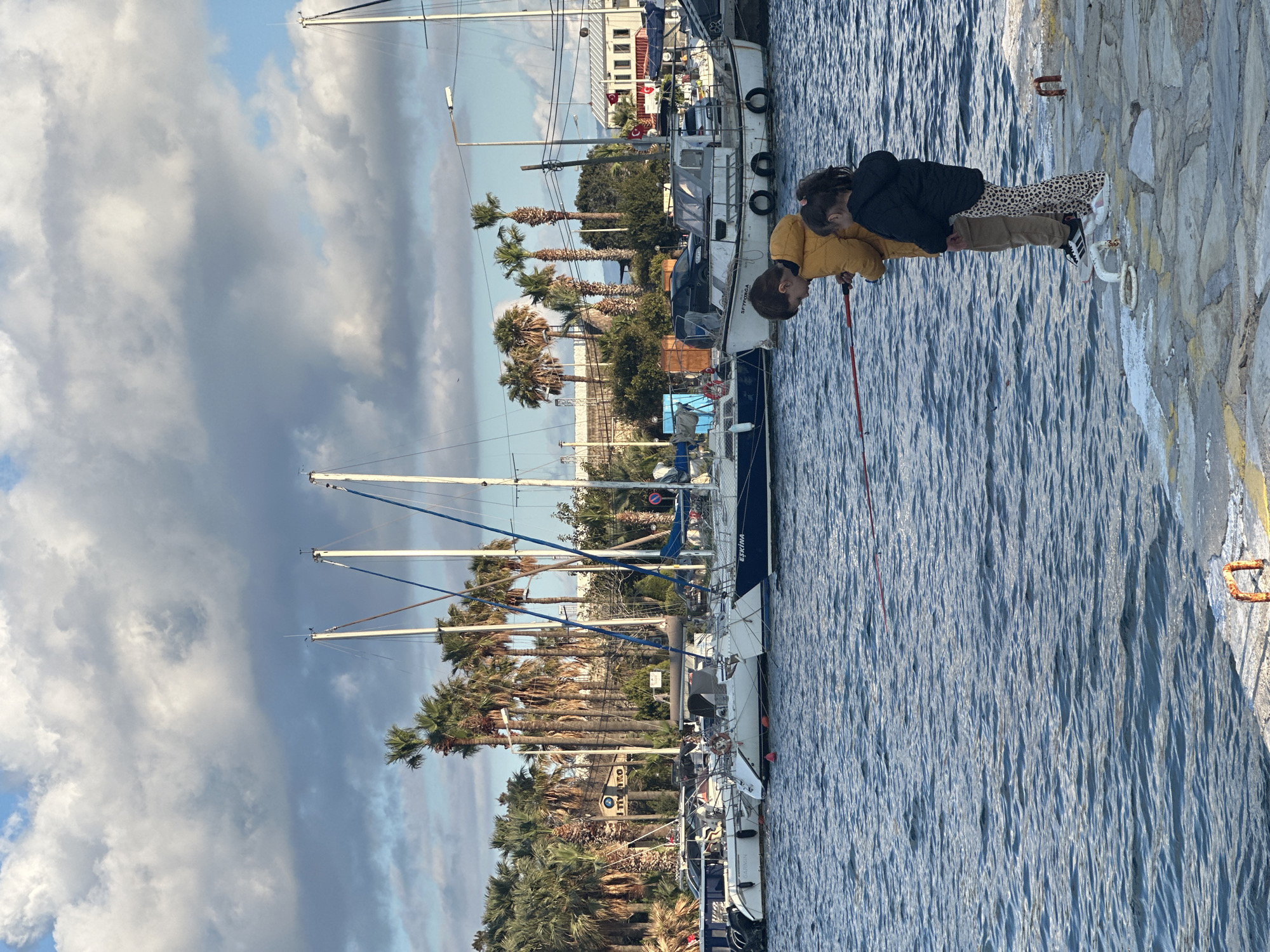 Two individuals, possibly children, are standing on a stone quay near the water's edge. One is wearing a yellow jacket and the other is in a black jacket with animal print pants. A long red object is visible in the hands of the individual in the yellow jacket.
The scene features several sailboats moored along the quay. Palm trees line the waterfront and in the background are buildings. The sky is partly cloudy. The location is Datça, Turkey, evidenced by Turkish flags on boats and the presence of distinctive local architectural styles. The water appears to be a harbor.