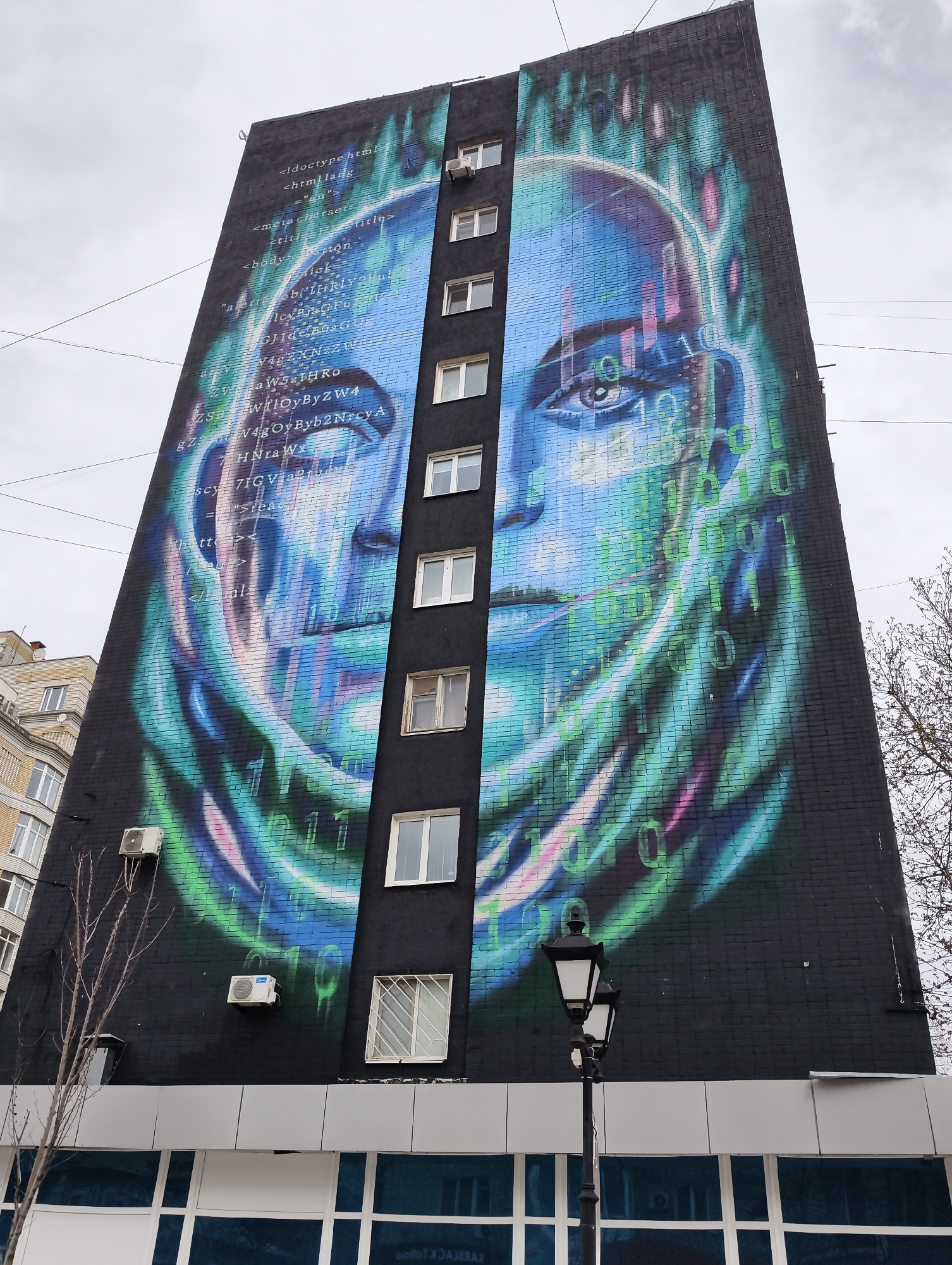 The image captures a striking mural on the side of a building in Saratov, Russia. The artwork depicts a blue-toned face with the eyes being prominent, intertwined with streams of binary code and fragments of HTML coding. The mural stretches vertically along the building's façade. The building itself is brick and has windows. On the lower portion of the building, below the mural, is a facade with white and clear panels. A black lamppost is in front of the building. The sky is overcast, and bare branches of a tree are visible on the right side of the image. Another building can be seen next to the building with the mural. There are also air conditioning units affixed to the building's exterior beneath the mural.