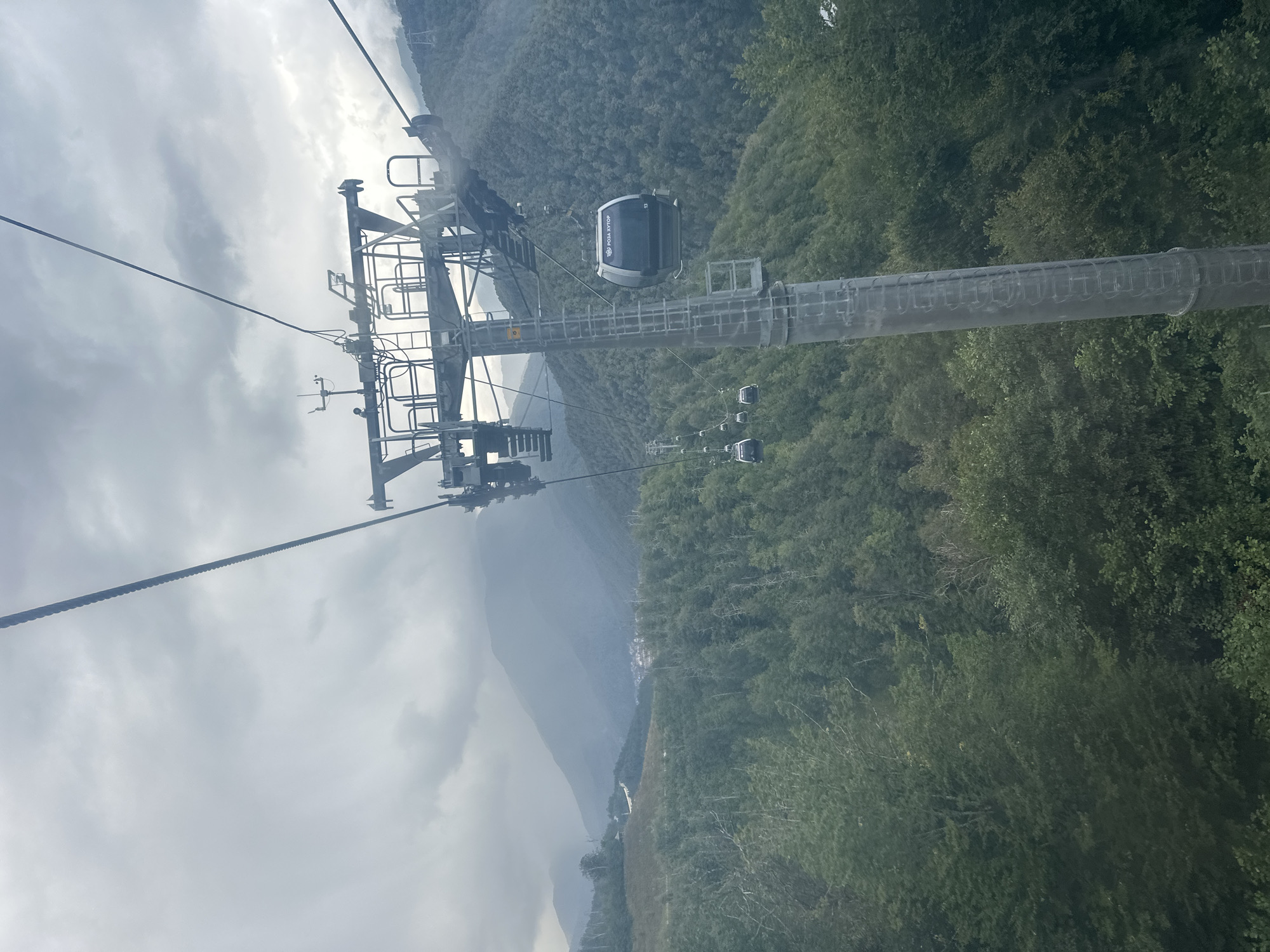 The image captures a view from a cable car ascending a mountain, likely in Estosadok, Russia, given the visible text on one of the cars. In the foreground, a massive pylon of the cable car system dominates the right side of the frame, with a cable extending diagonally upwards into the overcast sky. Several cable car cabins are visible; one prominent dark cabin, marked with the Cyrillic text "РОЗА ХУТОР" (Rosa Khutor) and the number "13," is positioned near the pylon. Further down the cable line, more cabins are spaced out, descending towards the valley. The surrounding environment is a lush, densely forested mountainside, with layers of green trees stretching towards the distant, hazy mountain peaks. The sky is heavily clouded, suggesting an overcast or potentially rainy day.