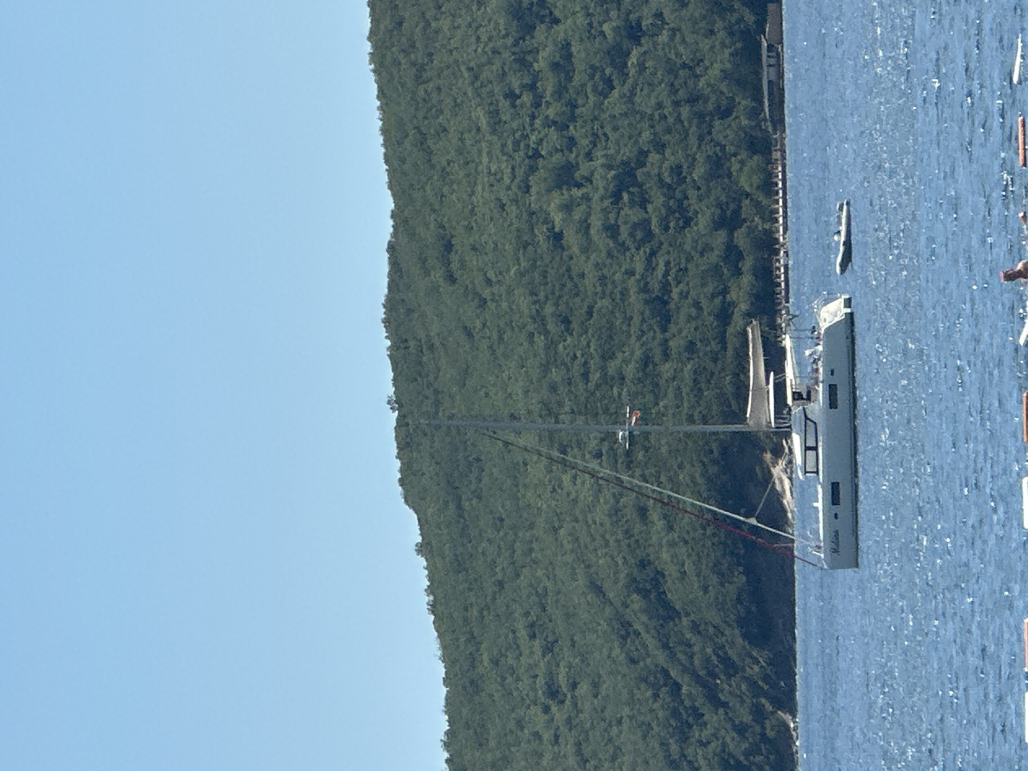 The image depicts a scenic view of a bay with a lush, green, tree-covered mountain dominating the left side of the frame. The mountain slopes steeply towards the water, creating a dramatic backdrop. The water is a deep blue, with a slightly choppy surface, suggesting a gentle breeze.
In the water, several objects are visible. A large, modern sailboat, predominantly white and grey, is positioned centrally. It has a tall mast and appears to be stationary or moving slowly. Further in the distance, a smaller white boat is also visible. Along the edge of the mountain, a pier or boardwalk stretches into the water. In the far right of the image, there are some orange buoys.
The sky is a clear, light blue, indicating a sunny day with good visibility. The light suggests it is daytime, likely mid-morning or afternoon. The scene appears to be a peaceful and picturesque location, possibly a popular tourist destination, given the presence of the sailboat and buoys. The image seems to be taken from a higher vantage point, looking down towards the bay.
There is faint text visible on the side of the large sailboat, which appears to read "Medusa". The overall impression is one of natural beauty and serene maritime activity.
