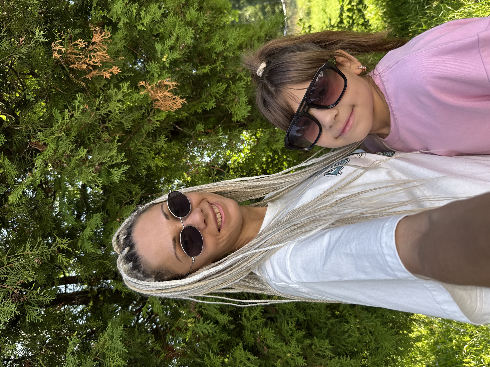 In Rusanovo, Russia, a woman with blonde box braids and oval sunglasses poses for a selfie. She is wearing a white t-shirt. To her right, a young girl with a ponytail and black aviator sunglasses is leaning into her. The girl is wearing a pink long-sleeved shirt. They are standing in front of a large green tree with some brown, dry branches. The background is out of focus, showing green grass and more trees.