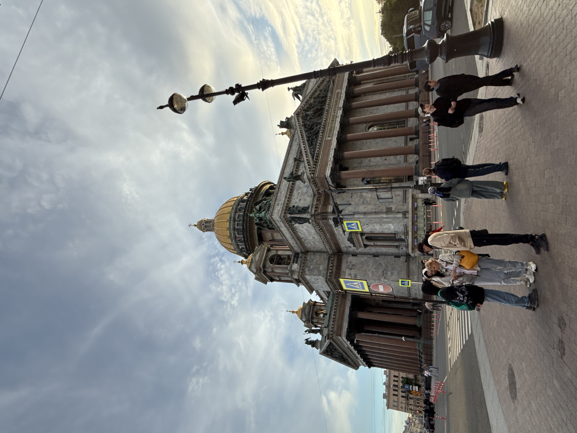 The image depicts a street scene in Saint Petersburg, Russia, with a prominent view of Saint Isaac's Cathedral. The cathedral, a grand architectural marvel with a golden dome, dominates the background. Several people are visible standing on the sidewalk, appearing to be tourists or locals. They are dressed in casual attire, suggesting an outdoor excursion. One group of people seems to be posing for a photo or observing the surroundings.
The street is paved with cobblestones, and a lamppost with multiple globes stands near the people. The sky is partly cloudy, with streaks of white clouds against a pale blue expanse, hinting at either early morning or late afternoon. The lighting suggests a moderate amount of daylight.
Notable details include the architectural grandeur of the cathedral, the presence of pedestrian crossing signs (one with a yellow background and a silhouette of a pedestrian, another with a red circle and a diagonal line), and the overall urban environment. The general atmosphere appears relaxed and observational, with no indication of any specific event or urgent activity. The orientation of the photo is on its side, making it appear vertical.