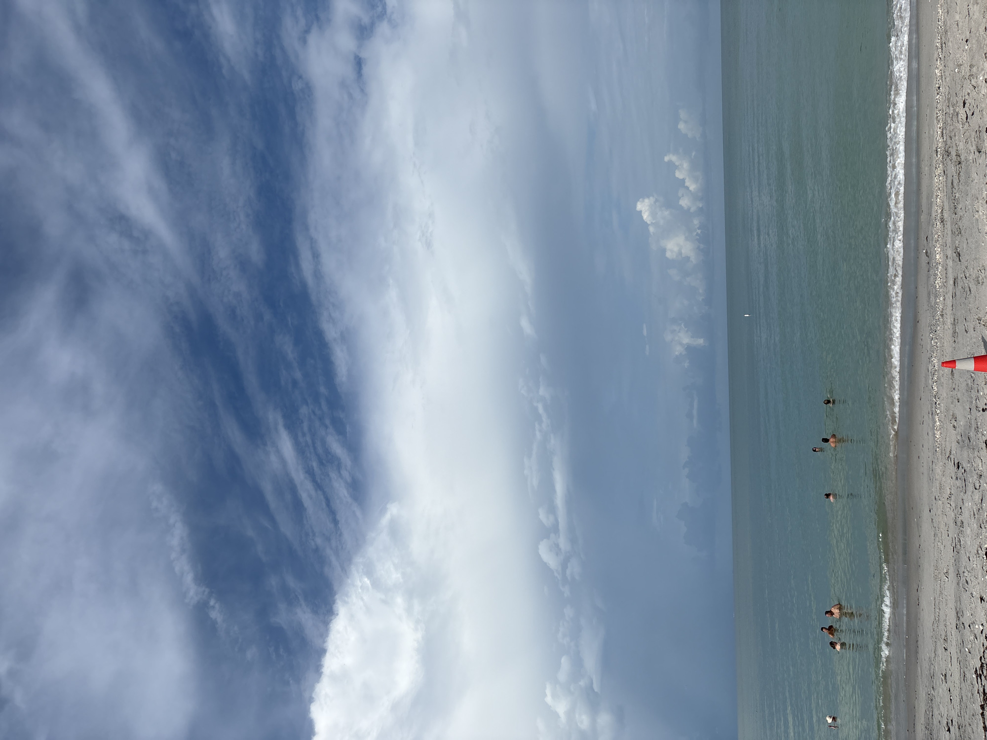This image depicts a wide shot of a beach scene in Englewood, United States. The ocean, with calm, turquoise water and gentle waves breaking on the shore, occupies the majority of the frame. The sky above is dramatic, filled with a mix of dark, stormy clouds and wispy white clouds against a blue background. Several people are visible in the water, appearing to be swimming or wading. On the sandy beach in the foreground, a bright orange traffic cone is positioned. The overall impression is one of a slightly overcast but warm day at the beach.