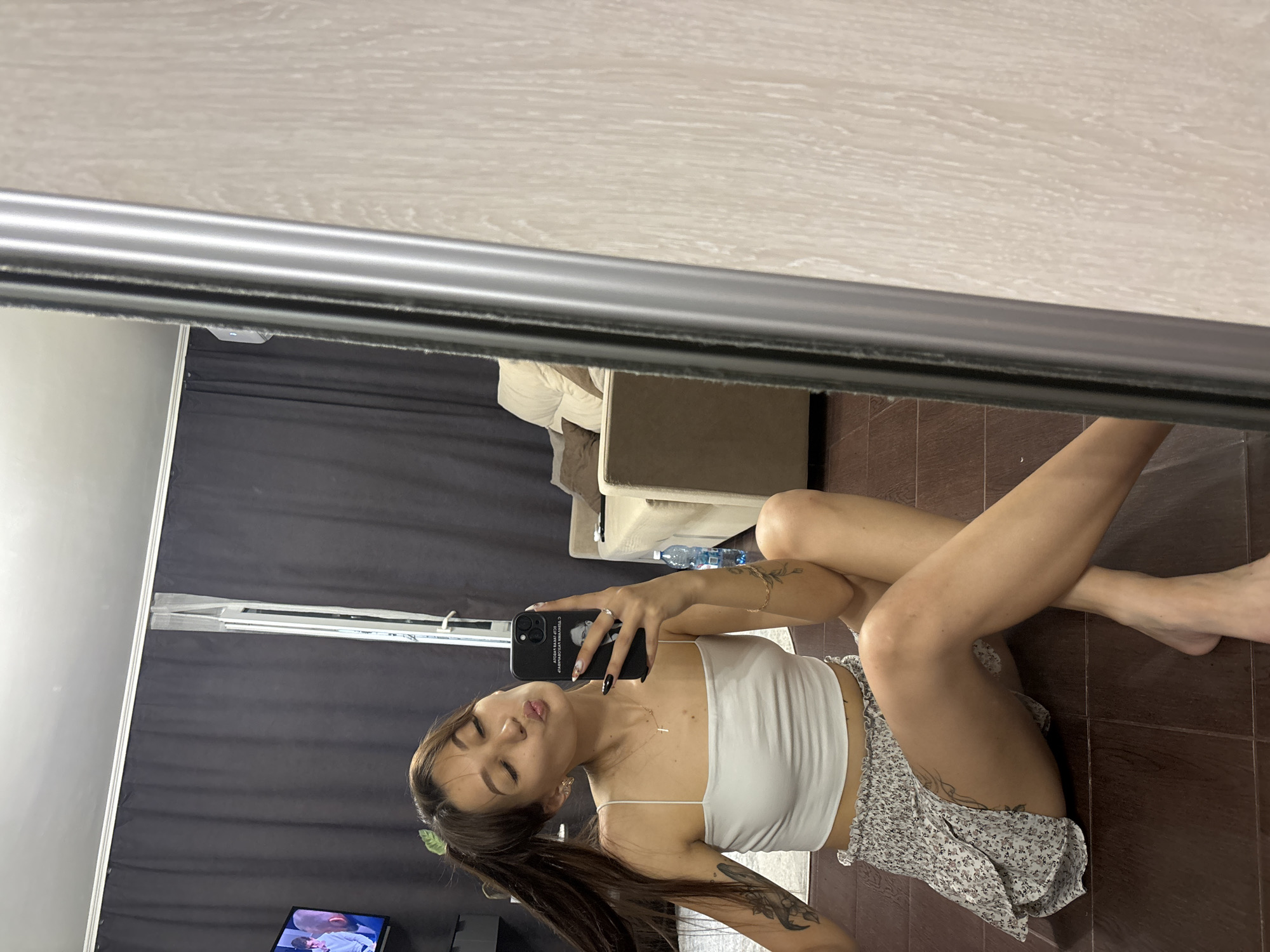 The image shows a young woman taking a selfie in a dimly lit room. She is sitting on a wooden floor with her legs crossed. She is wearing a white tank top and a floral print skirt. She has long dark hair tied in a ponytail and is wearing minimal jewelry. Her face is turned towards the phone, and she is making a kissing face. Tattoos are visible on her arms and legs. In the background, a large mirror reflects the room, showing a sofa, a bottle of water, and a TV screen displaying an image. The room appears to be a bedroom or living area, with dark curtains and a light-colored wall. The overall mood is casual and playful. The scene seems to be indoors, with no indication of the time of day or weather conditions.