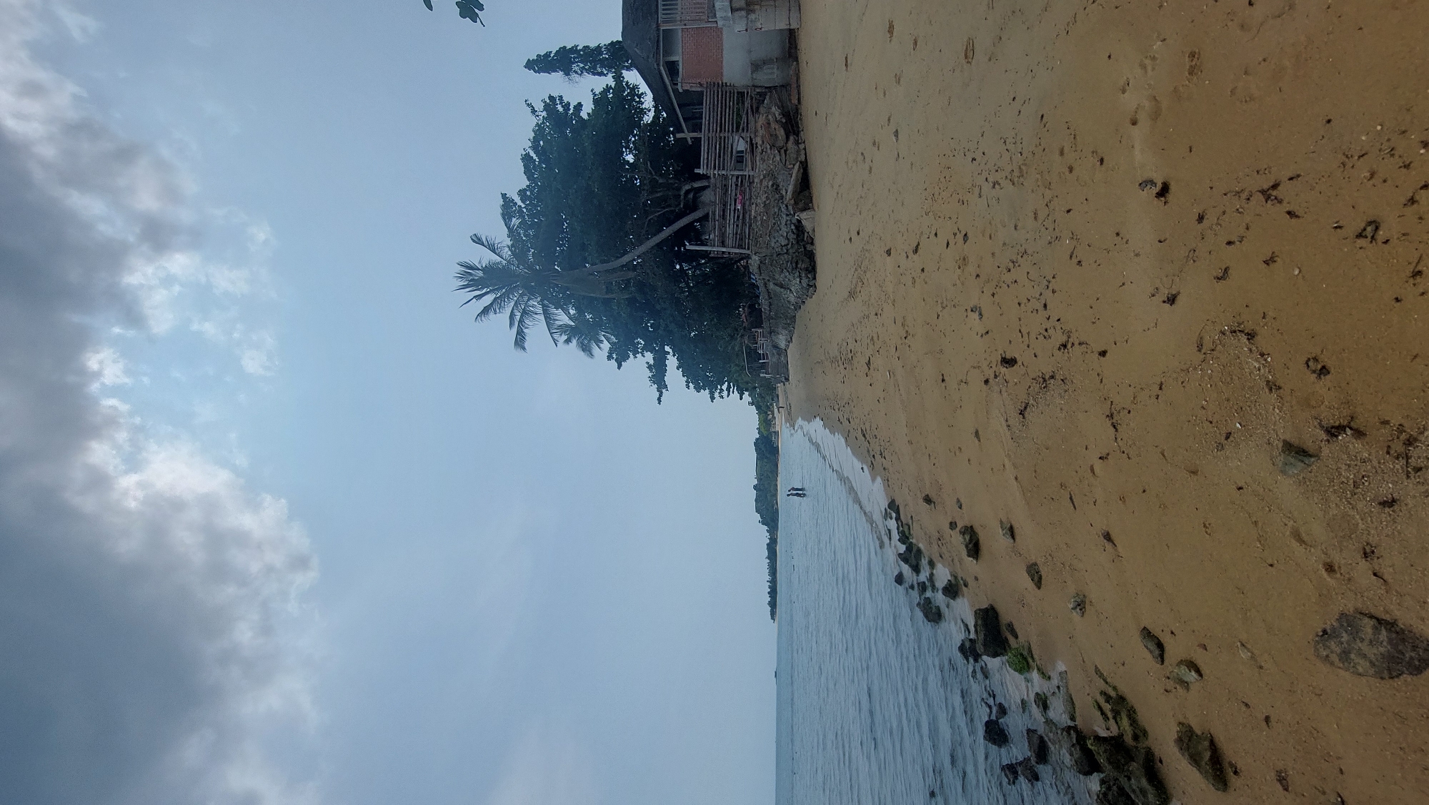 The image captures a beach scene in Baan Mae Nam, Thailand. The sky is predominantly overcast with cloudy formations. The beach is characterized by light brown sand with scattered debris and rock formations bordering the water's edge. A building, possibly a small structure or house, is situated behind a dense cluster of trees, including a palm tree. Two figures are present in the water, likely walking or standing. The water appears calm with gentle ripples.