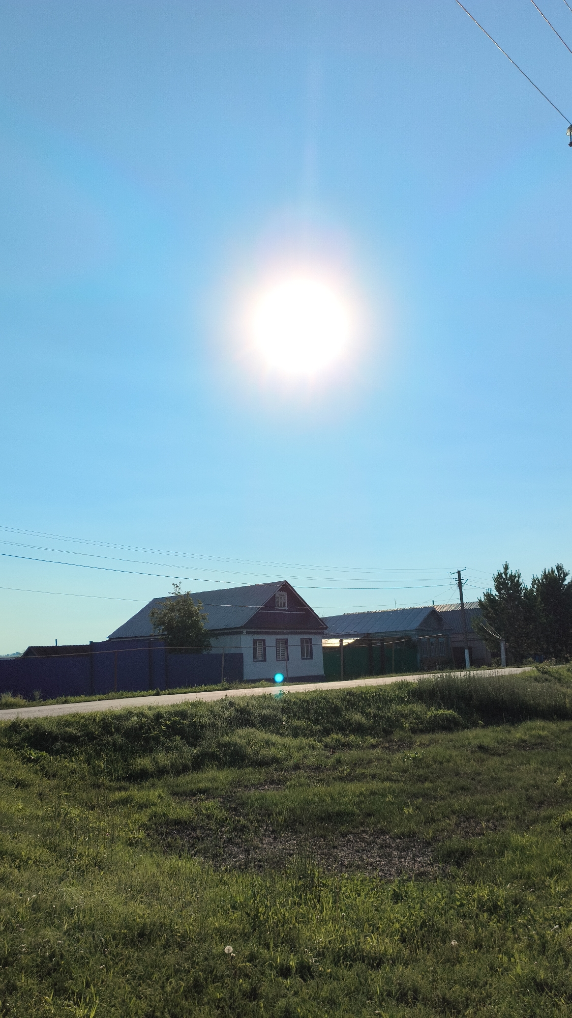 The image depicts a bright, sunny day in the village of Alkin, Russia. The sky is a clear, vibrant blue, dominated by a large, intense sun near the top of the frame, casting a bright glare. In the middle ground, a long, low-rise white house with a dark roof stands prominently, flanked by a dark blue fence and a green fence. Adjacent to this house, another similar structure is visible. A road or path runs horizontally across the scene, separating the foreground from the houses. The foreground is a lush, green grassy field, dotted with sparse white dandelion seed heads. Overhead, electrical wires stretch across the sky, supported by utility poles visible in the distance. The overall impression is a rural, tranquil scene bathed in strong sunlight.