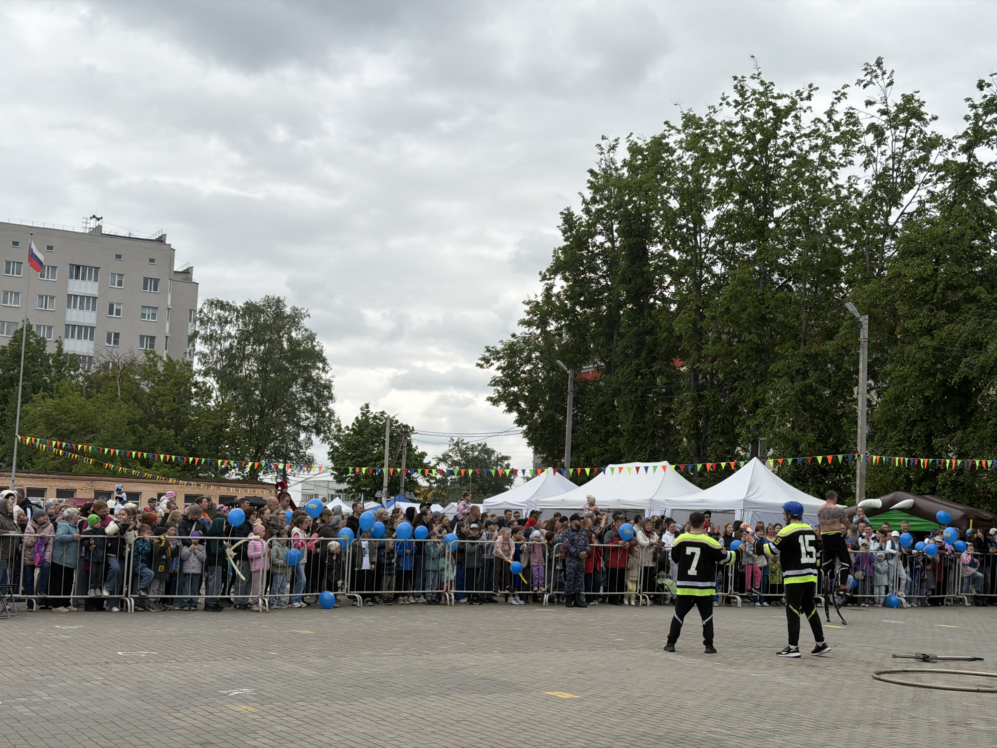 A medium-shot shows a crowd behind a low fence facing away from the camera in Staraya, Russia. In the foreground, two performers wearing black and yellow jerseys with the numbers "7" and "15" are preparing to perform for the crowd. The crowd is composed of people of all ages, some holding blue balloons. In the background, there are white tents, green trees, and a gray building with a Russian flag. The event is taking place on a paved surface. The sky is overcast.