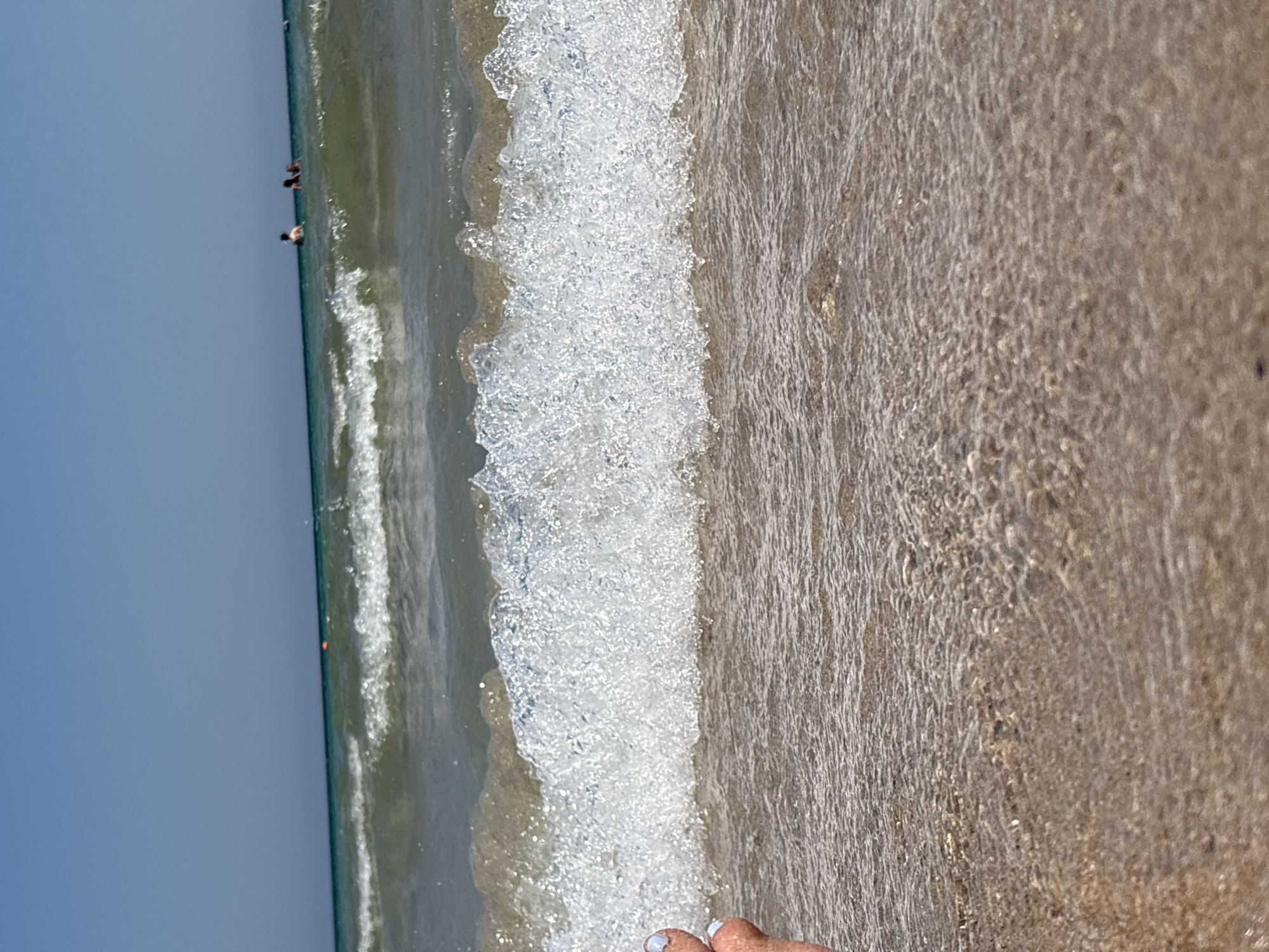 The image shows a close-up view of a gentle wave rolling onto a sandy beach. The water is clear and shallow, with sunlight reflecting off the sandy bottom. In the background, several people are swimming in the ocean. The sky is clear and blue. The location is a beach in Primorsko, Bulgaria.