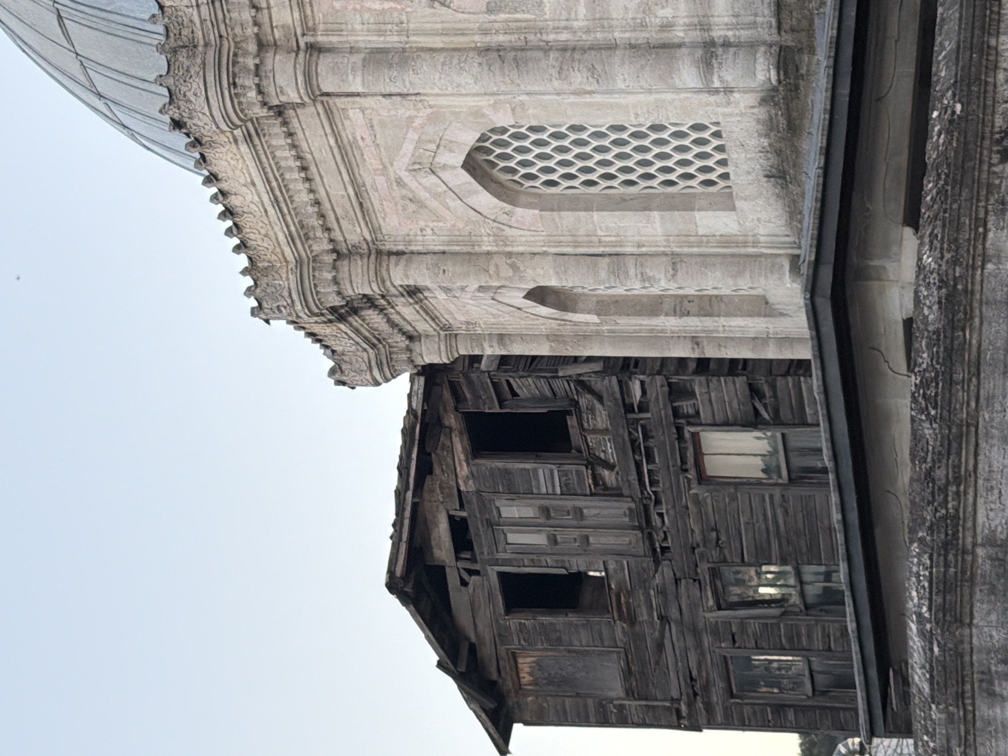 This image depicts a stark contrast between a dilapidated wooden structure and an ornate, classical building with a domed roof. The wooden building, positioned on the left, appears abandoned and in severe disrepair, with weathered planks, broken windows, and a sagging roofline. Its dark, aged wood suggests a long history and neglect.
To the right, the classical building stands in stark contrast, showcasing intricate architectural details. It features stone construction with decorative carvings along the upper facade, fluted columns, and arched windows with lattice-like grilles. A portion of its dome, covered in what appears to be metal sheeting, is visible at the top of the frame.
The two structures are positioned adjacent to each other, highlighting a juxtaposition of decay and preservation. The environment appears to be outdoors, with a pale sky suggesting either early morning or late afternoon light. The presence of other structures or vegetation is minimal, with only a hint of greenery on the far left, possibly suggesting a tree line or distant landscape. The image does not contain any people or visible actions. The scene is static, focusing solely on the architectural elements and their contrasting states of condition.
The architectural styles and the presence of a domed building strongly suggest this image was taken in Eyüpsultan, Turkey, a district known for its historical mosques, tombs, and traditional wooden Ottoman houses. The contrast between the decaying Ottoman wooden house and the more enduring stone structure is a common sight in historical urban areas undergoing development or preservation.