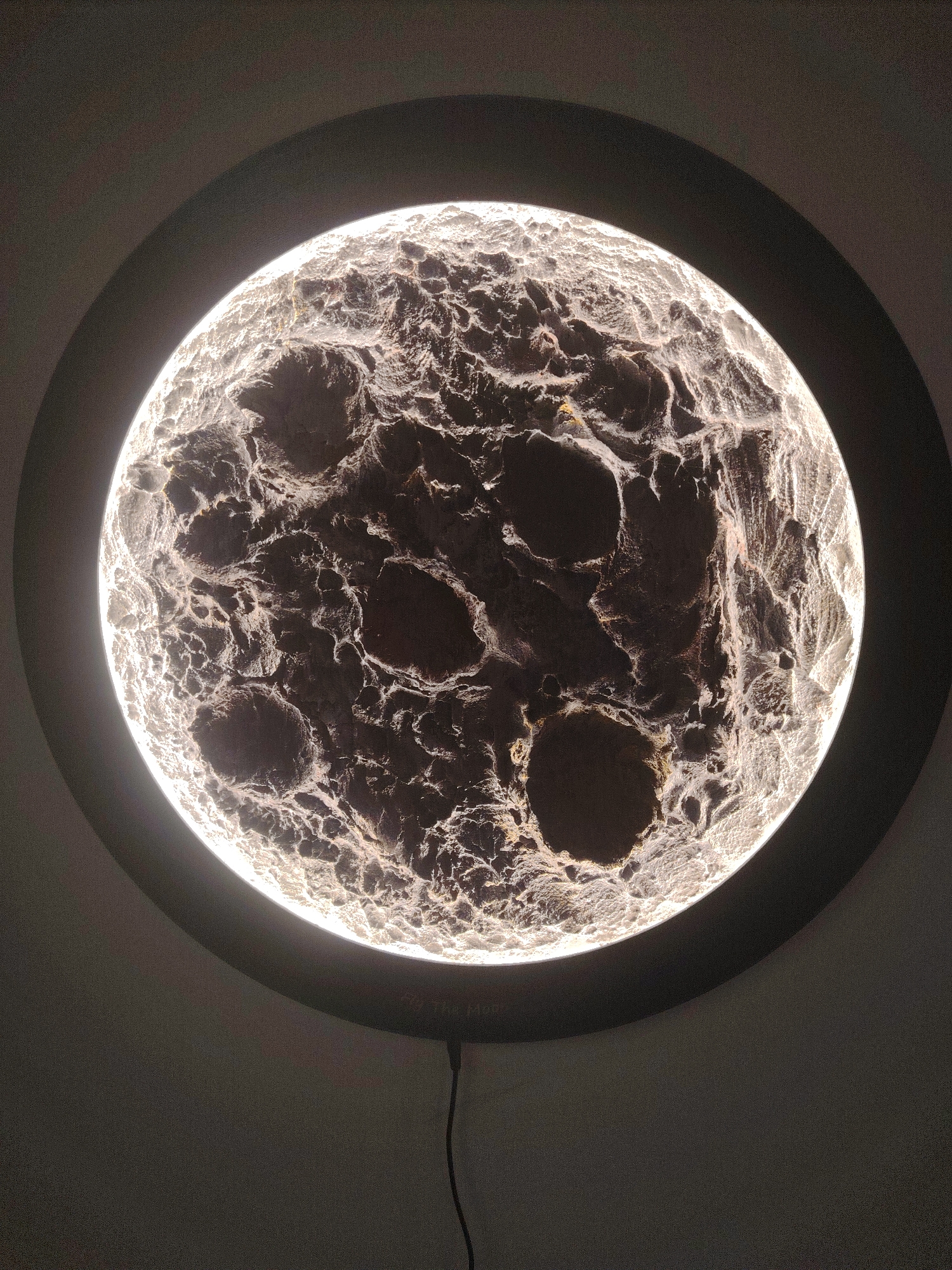 The media file displays a circular light fixture or art piece, designed to resemble the moon's surface, set against a plain, dark-grey wall. The central disc is intensely illuminated from within, highlighting the intricate, textured topography of craters, ridges, and valleys. This strong backlighting creates a striking contrast, making the elevated areas appear bright white and the deeper depressions dark, enhancing the three-dimensional effect. A clear halo of light emanates from the edge of the illuminated surface, bordered by a dark, circular frame.
A black power cord extends downwards from the bottom edge of the frame, indicating an electrical connection. The background is an unadorned wall, suggesting an indoor setting. No activity or event is taking place; the image is a still photograph of the object itself. The ambient lighting conditions appear dim, allowing the glowing object to be the primary light source in the scene. Faint white text, which appears to read "Art Orb", is visible on the lower section of the dark circular frame, slightly above where the cord emerges. The location context provided for this image is Huntingdonshire, United Kingdom.