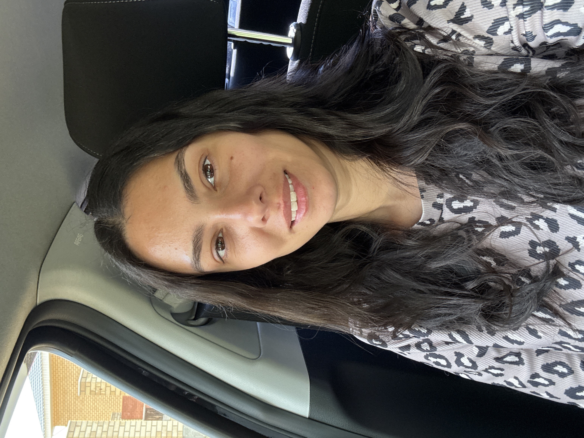 A woman with long, dark, wavy hair and brown eyes is shown from the chest up, seated in the passenger seat of a car. She is wearing a light-colored long-sleeved shirt with a black and white leopard print. The woman is smiling and looking directly at the camera. The interior of the car is visible, including a black headrest and part of the car's door frame. Outside the car window, a building with yellow brick siding and a light-colored roof is visible, suggesting an urban or suburban setting. The presence of a car and a building implies a location within a populated area. Given the request for specificity related to Chaikovsky, Russia, the scene is likely situated within the city or its immediate surroundings, potentially a residential area or a street. The lighting indicates it is daytime.
