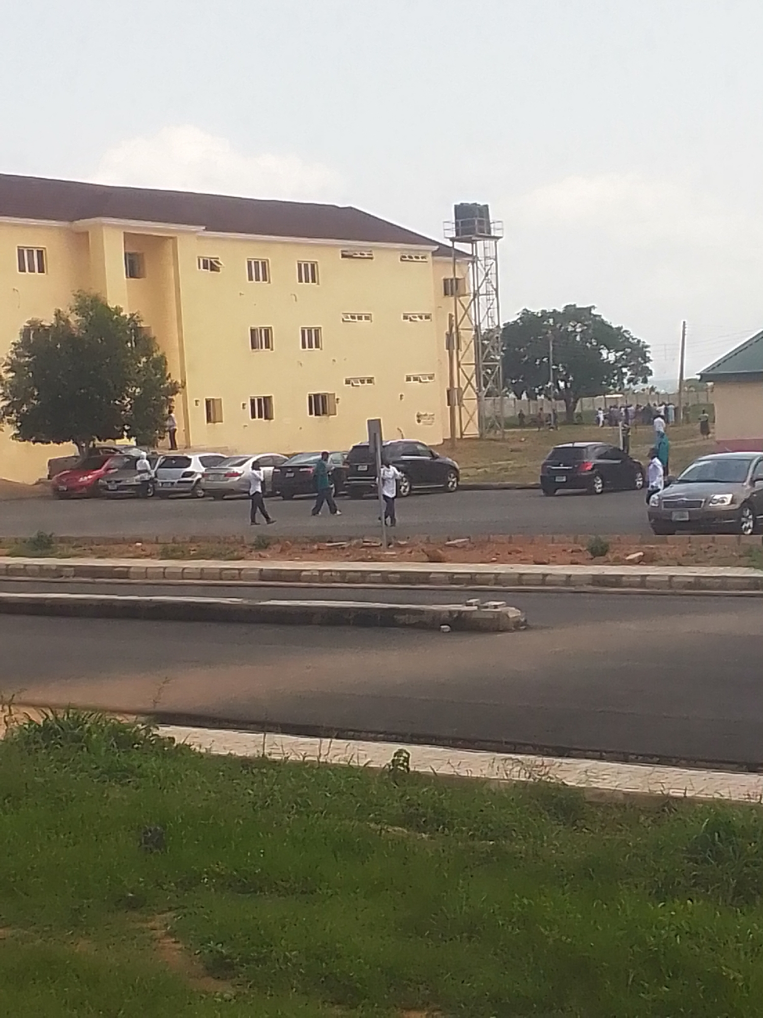 The image depicts an outdoor scene in Kachia, Nigeria, likely during daylight hours given the bright ambient light. A long, light yellow, multi-story building with a dark roof dominates the background. The building features numerous rectangular windows. To the left of the building, a large, leafy tree provides shade. Adjacent to the building is a parking area with several cars, including a red sedan, a white hatchback, and a dark-colored SUV.
In the foreground, a paved road curves into the frame. Several people are walking or standing in the parking area and near the road. Some appear to be students or residents of the building. A tall, metallic water tower stands to the right of the building. The weather appears to be overcast or hazy, suggesting a diffused light. The scene conveys a sense of everyday activity, possibly on a university campus or a residential complex. No visible text is present in the image.
