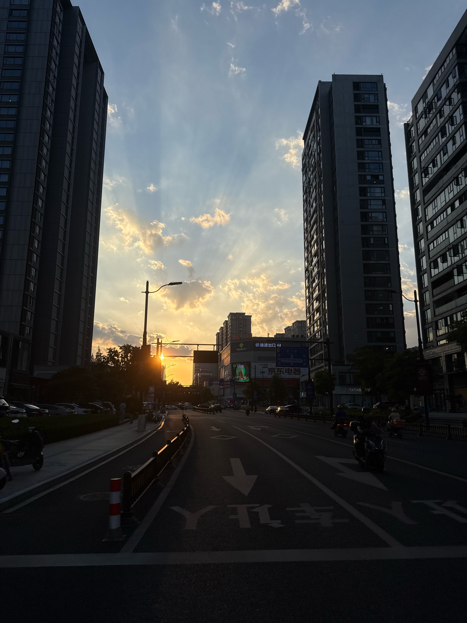 The image shows a street scene in Yiwu, China, during sunset. Tall, modern buildings line both sides of the street, casting long shadows. The sky is dramatic, with rays of sunlight breaking through the clouds, illuminating the lower portions of the clouds with a warm glow. The sun itself is visible, a bright orb peeking through the cloud cover, positioned low on the horizon between two buildings.
The street is paved and appears to be a multi-lane road with traffic markings, including directional arrows. Several vehicles, including cars and motorcycles, are on the road, some in motion and others parked along the side. A few pedestrians can be seen walking on the sidewalk. A traffic light pole stands near the center of the image, and various signs and advertisements are visible on the buildings. The overall impression is of an urban environment at dusk, with the natural beauty of the sunset contrasting with the man-made structures.