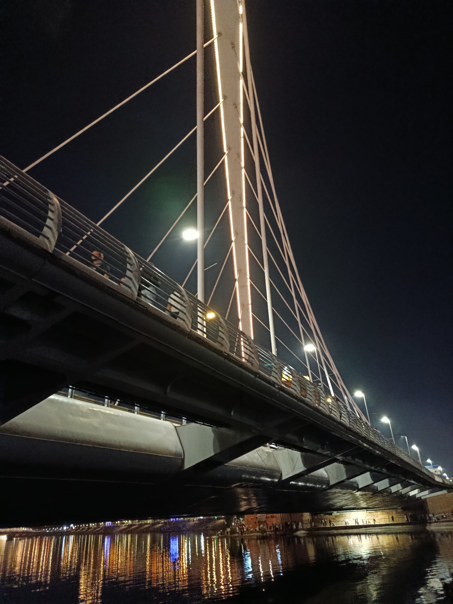 This low-angle, night-time photograph captures the modern architecture of the Pedestrian Bridge over the Ishim River in Astana, Kazakhstan. The bridge features a prominent pylon structure from which numerous white cables extend to support the walkway, which is lined with a metal railing and illuminated by warm-toned lights. Below the pedestrian level, the underside of the bridge is visible, a dark, industrial structure. The river below reflects the vibrant lights of the city, creating a shimmering, colorful pattern on the dark water. Several figures are faintly visible walking on the bridge's pedestrian path and along the embankment below. The overall atmosphere is one of urban nighttime beauty and modern engineering.