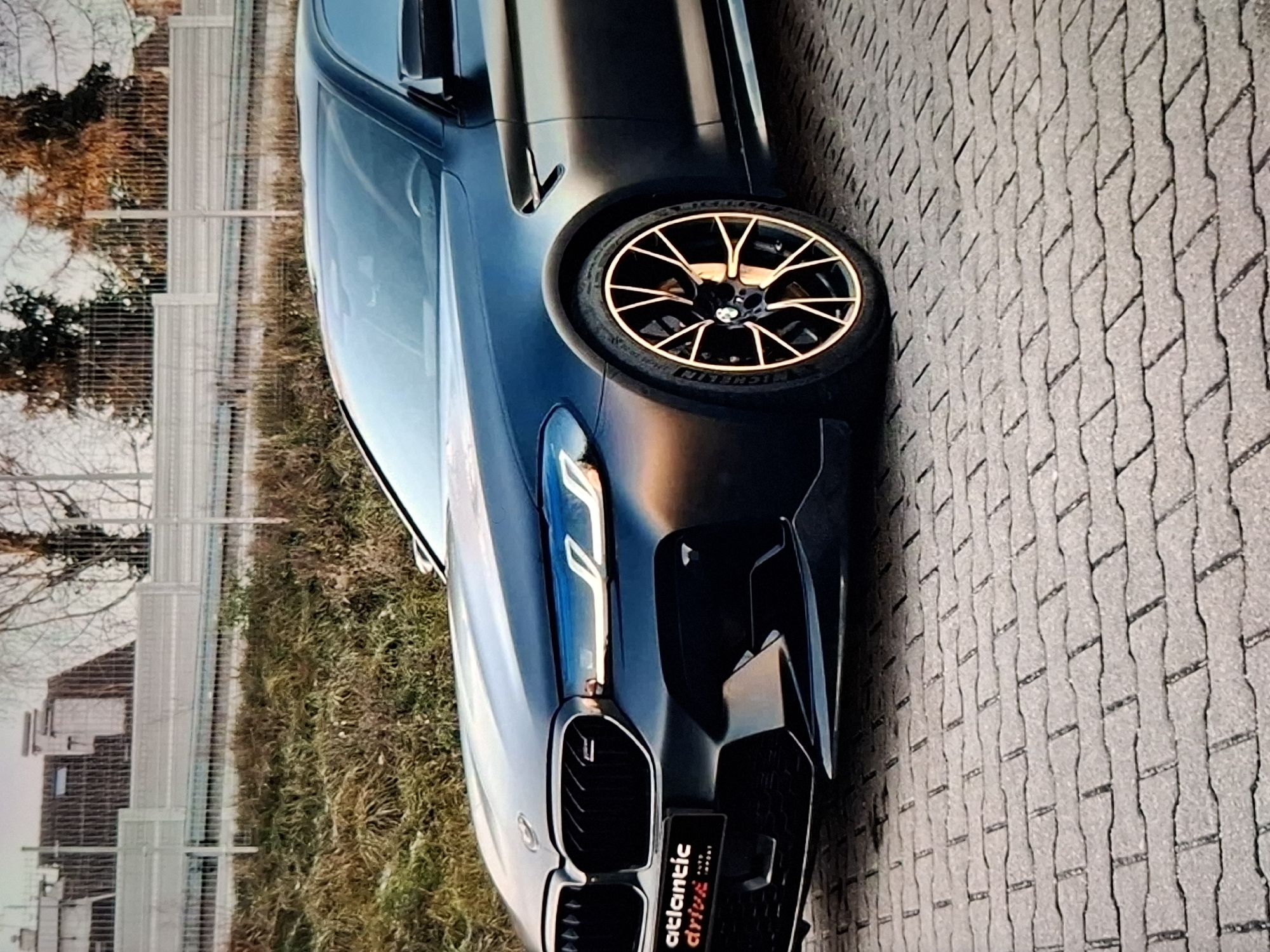 The image shows a close-up, low-angle view of a black BMW M5 sedan parked on a brick-paved surface. The car's front grille, headlights, and a portion of the hood are visible. The headlights have a distinctive daytime running light signature. The car has aftermarket wheels with a copper or bronze finish. A small black badge with white text "ATLANTIC DRIVE" is affixed to the lower left of the grille. In the background, a dry, grassy embankment is visible behind a metal fence. Beyond the embankment, there are bare trees and a building with a sloped roof. The lighting suggests it is either early morning or late afternoon. There are no people or actions depicted in the image. The scene is an outdoor setting, likely a parking area or roadside. The location is not Urus-Martan, Russia.