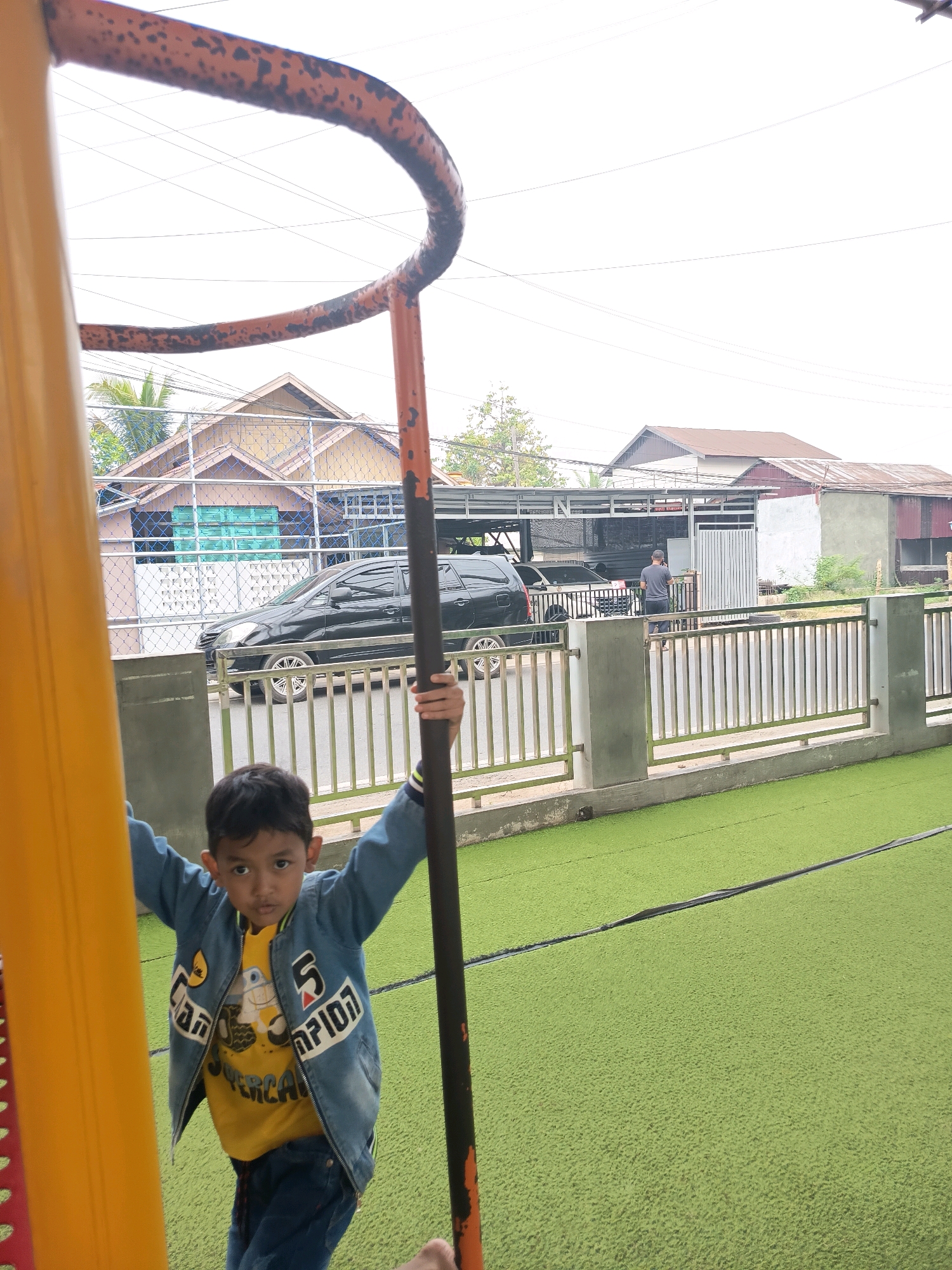A young boy of Southeast Asian descent, appearing to be between 5-7 years old, is captured in a low-angle shot from the perspective of a playground. He is wearing a yellow t-shirt with a cartoon character and a blue denim jacket adorned with white lettering. The boy is holding onto a vertical pole and a yellow structure, looking towards the camera with a neutral expression.
Behind the boy, a black car is parked on a road next to a green artificial turf surface. Beyond the road, there are houses with brown and grey roofs, some with visible windows and fences. A man in a grey t-shirt and dark pants can be seen in the background, standing near a building. The scene appears to be outdoors, with power lines visible against an overcast sky. The environment suggests an urban or suburban setting, potentially a playground or park area within Banjarmasin, Indonesia, given the architectural styles and the likely climate.