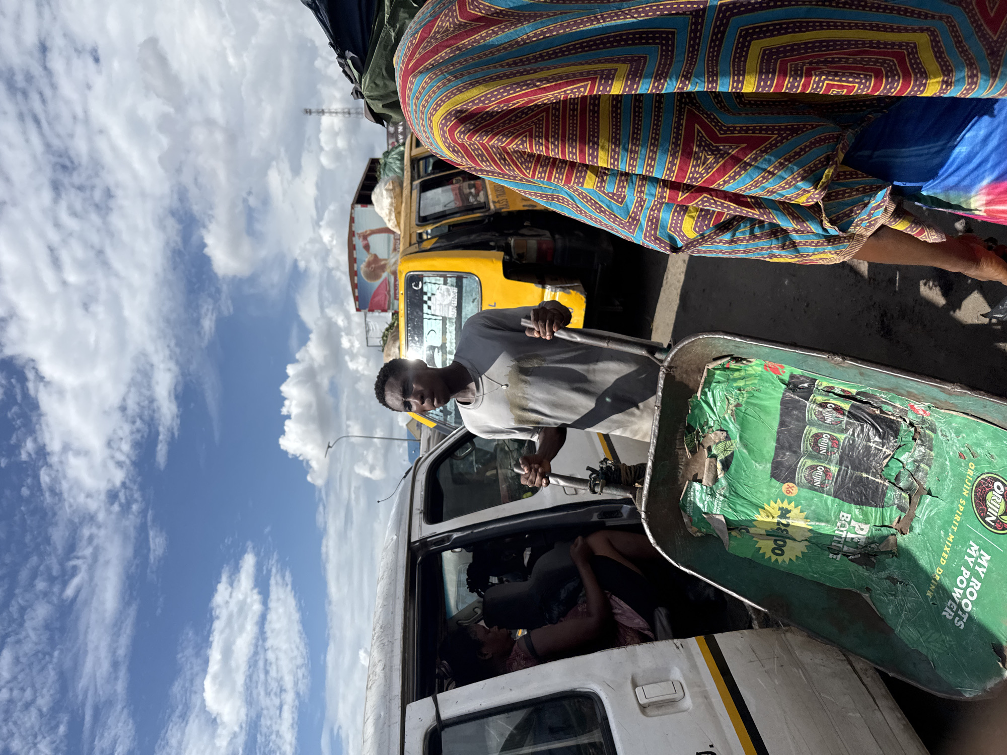 In the Fegge area of Nigeria, a congested street scene unfolds. A white passenger van sits with its door open, occupied by a woman reclined inside. A man stands outside, holding the handle of a green metal cart filled with packaging for "Origin" spirit mixed drinks. The sky is blue with scattered clouds overhead. In the background, a yellow taxi cab is visible, and a person wearing a colorful patterned garment stands in the foreground.