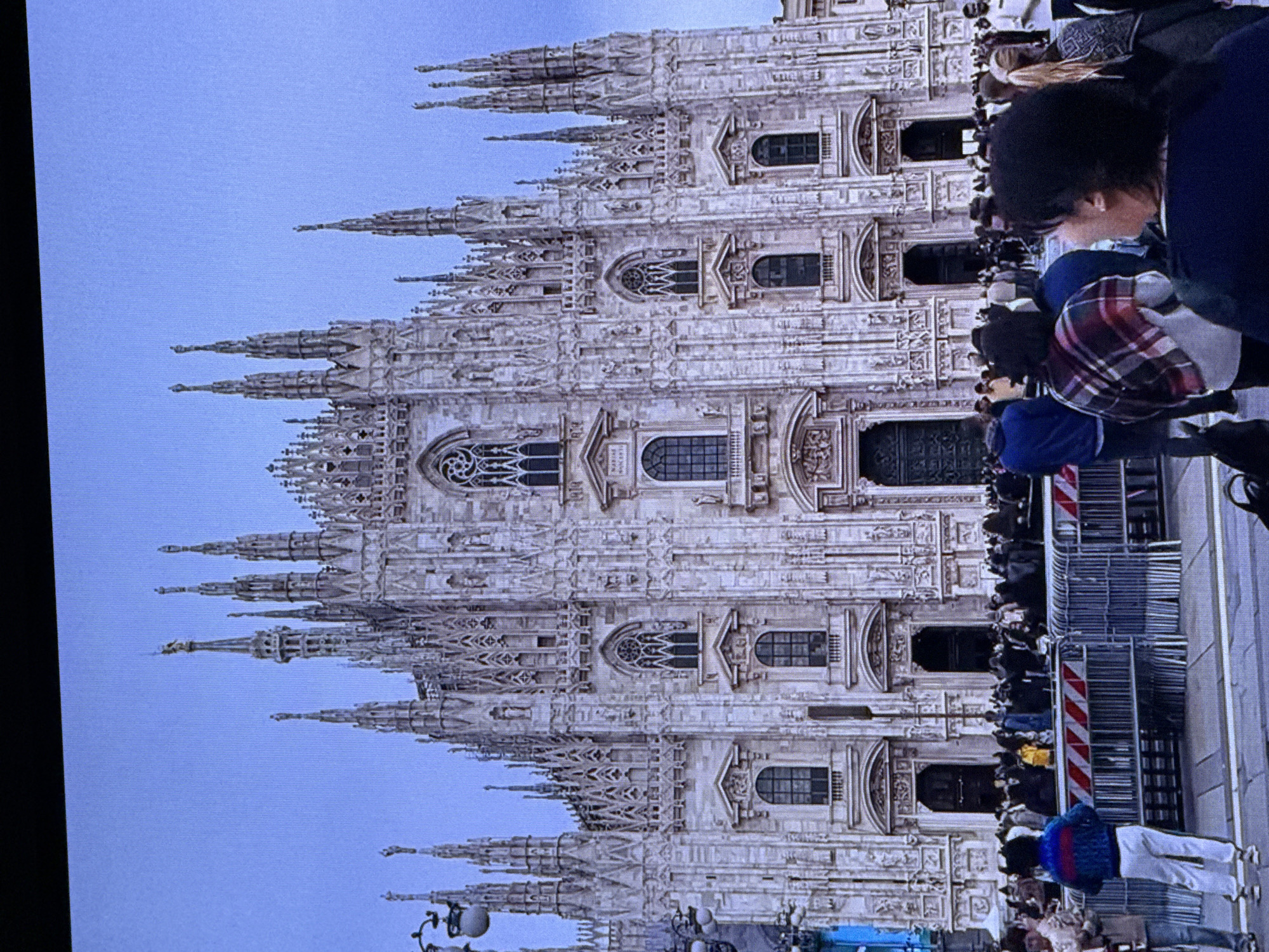 The image shows the Milan Cathedral, a historic Gothic cathedral in Milan, Italy, not Dubai, United Arab Emirates. A crowd of people is gathered in front of the cathedral, separated by metal barriers. The cathedral is a large, ornate building with many spires and intricate architectural details. The sky is a pale blue, indicating daylight. The people in the foreground are facing the cathedral, and some appear to be looking at their phones. The overall scene suggests a tourist or public gathering at a famous landmark.