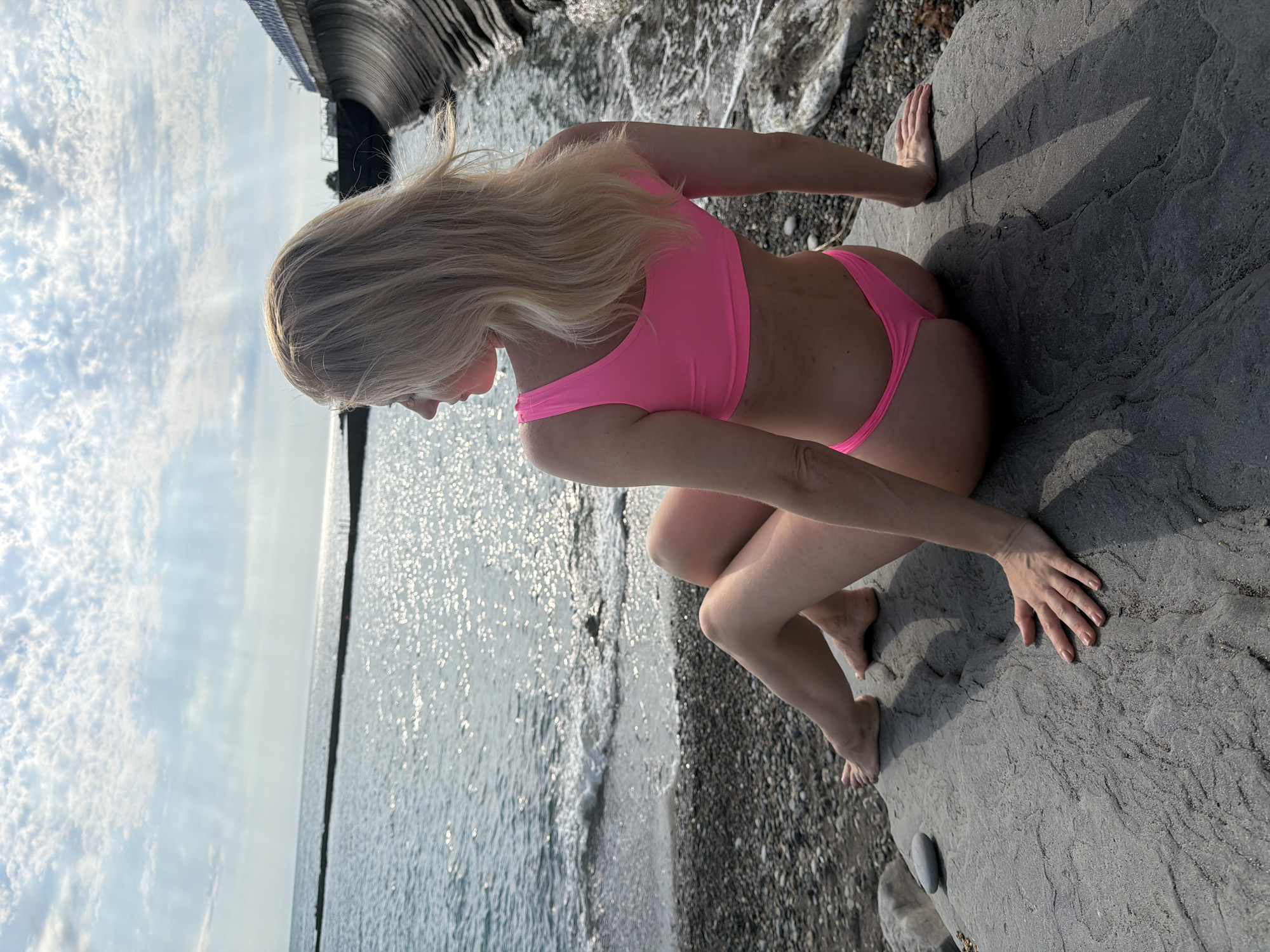 A blonde woman in a pink bikini sits on a large rock on a beach in Sochi, Russia, facing away from the camera and looking out at the sea. The sky is partly cloudy, with sunlight creating a shimmering effect on the water. The beach is covered in pebbles, and a concrete structure can be seen on the right side of the image. The woman's legs are crossed, and her hands rest on the rock. The tide is out, with gentle waves washing ashore.