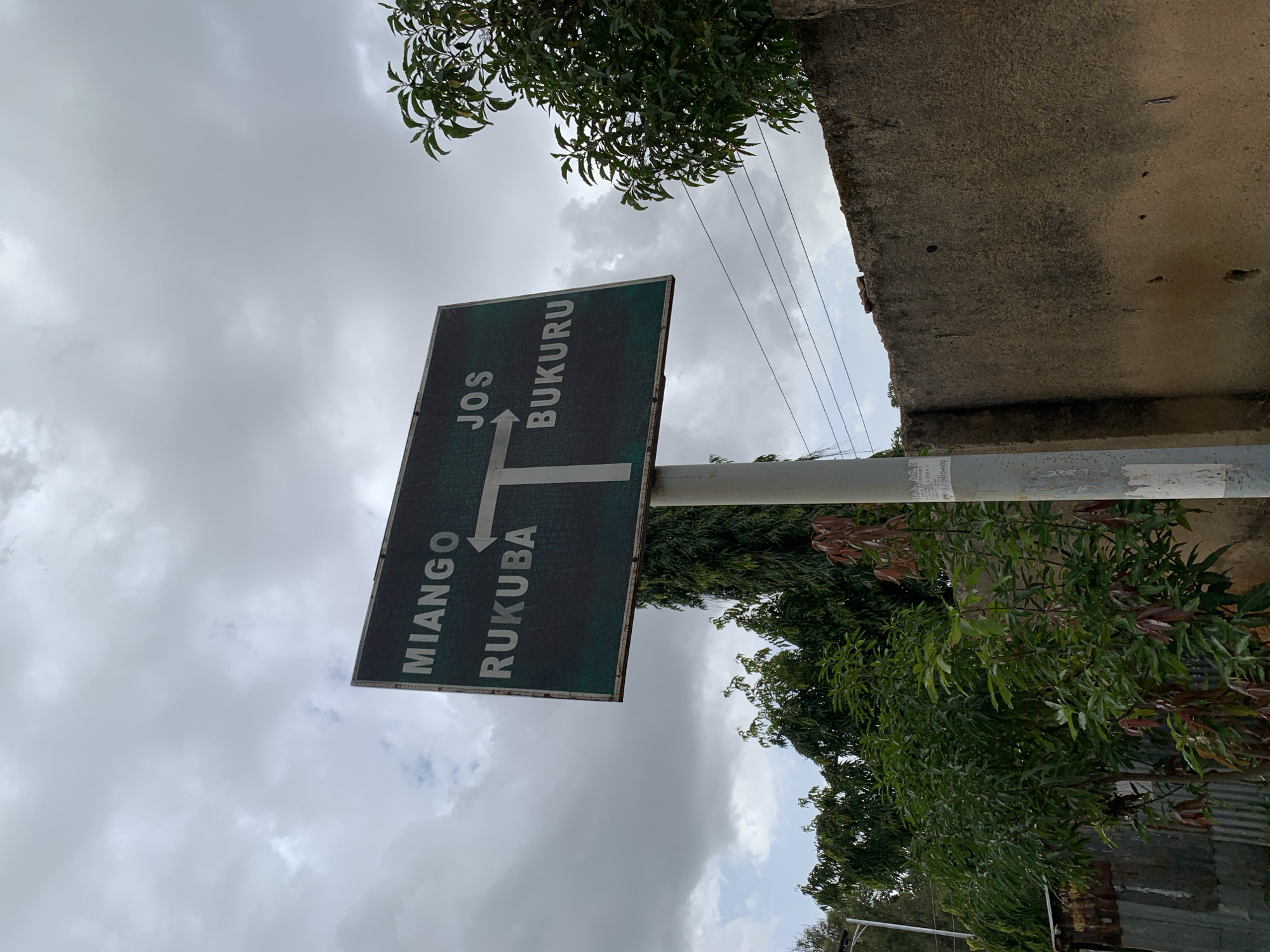 A green directional sign is mounted on a pole. The sign has two sections, each with white lettering. The left section indicates "MIANGO" and "RUKUBA" with an arrow pointing left. The right section indicates "JOS" and "BUKURU" with an arrow pointing right. The sign is positioned at a T-junction. In the background, there is lush green foliage and trees, a grey concrete wall, and a cloudy sky. A narrow metal fence is visible to the left of the sign. The overall scene suggests an outdoor location within a city, likely at a road intersection.