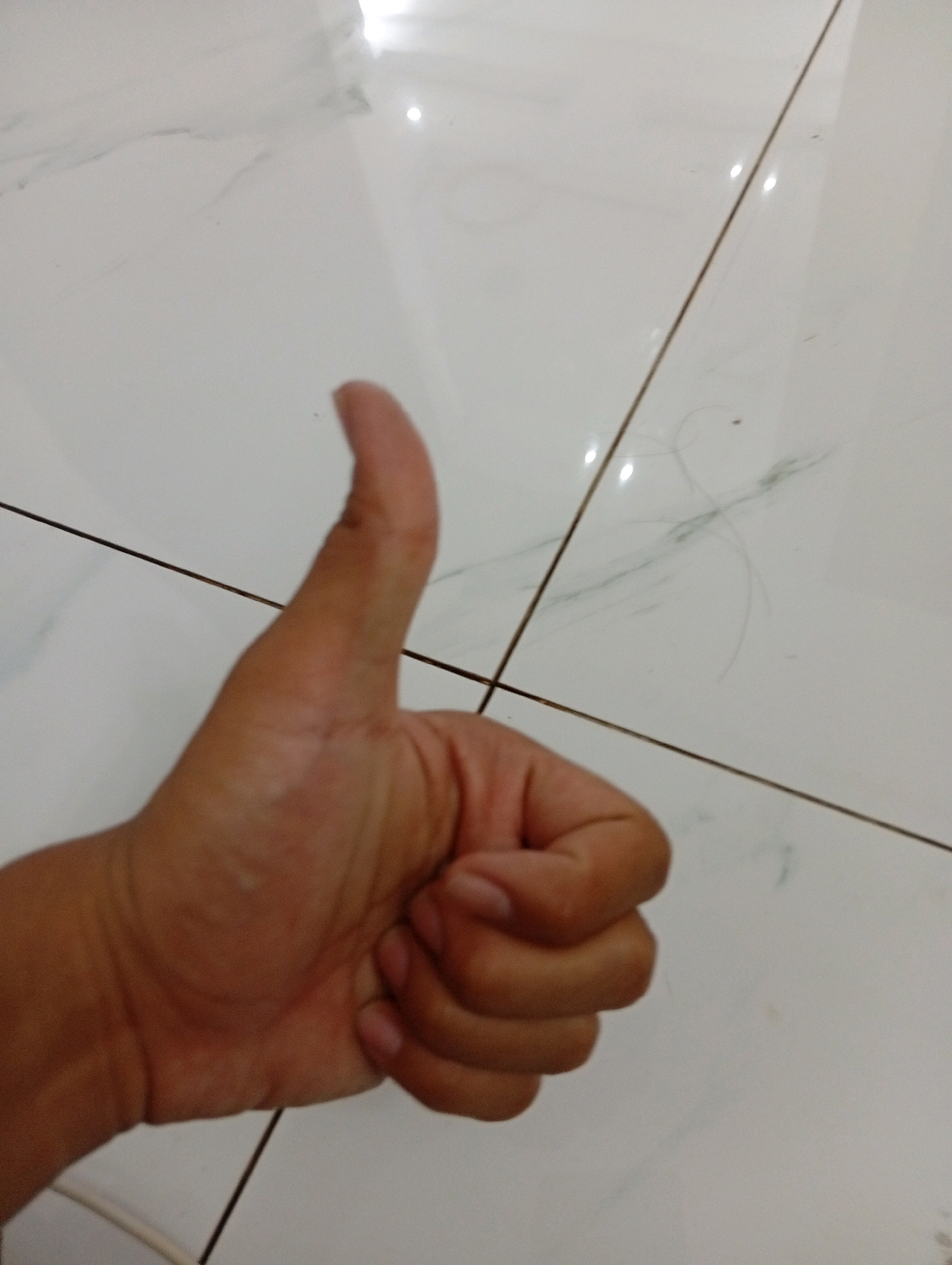 A hand is shown giving a thumbs-up gesture on a white tiled floor with visible grout lines. There are some strands of hair and faint green marks on the floor. The image is taken from a downward angle, focusing on the hand and the floor. The lighting appears to be from an overhead source, creating reflections on the tiles. The background consists of a tiled surface, likely a floor, with distinct grout lines creating a grid pattern.