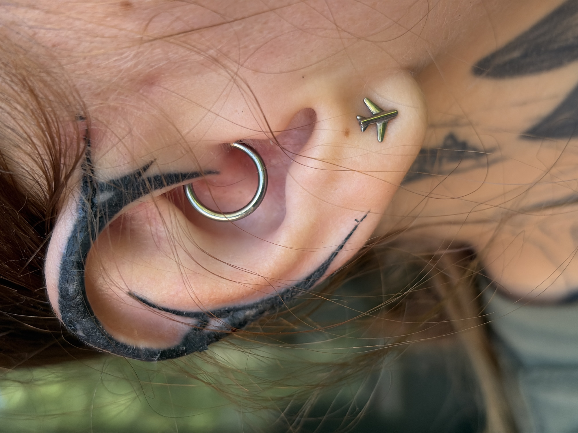 This is a close-up, eye-level, horizontal shot of a person's ear in an outdoor setting, likely in Chaikovsky, Russia. The ear features two piercings: a silver hoop piercing in the lobe and a small, silver stud earring shaped like an airplane in the upper part of the ear. The ear also has black tribal-style tattoos wrapping around the outer edge and extending towards the temple. Strands of light brown hair frame the ear, some falling across it. The background is softly blurred, with hints of green foliage suggesting a natural environment, possibly a park or garden. The lighting appears to be natural daylight. No text is visible in the image. The overall impression is one of personal adornment and a relaxed, possibly sunny, day.