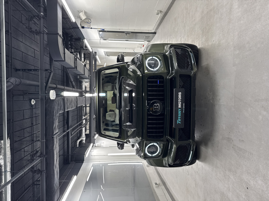 A dark green Brabus-modified Mercedes-Benz G-Class SUV is centrally positioned in an indoor facility, likely a luxury car showroom or detailing bay, located at Tiffany Motors, Aleje Jerozolimskie 200, 02-486 Warsaw, Poland. The vehicle faces the viewer directly, featuring a prominent "B" logo on its black grille, illuminated circular daytime running lights, a custom front bumper with mesh grilles, and a roof-mounted light bar. A black display plate on the lower front bumper reads "TIFFANY MOTORS" in blue text, with smaller white text "Tiffany Motors" underneath. The vehicle's interior shows light-colored seats.
The environment consists of a light gray concrete floor and white walls. To the left, a large reflective glass or panel partition is visible. To the right, a light-colored door with a vertical handle and what appears to be a wall-mounted air hose or cleaning tool are present. The ceiling is an exposed industrial type, painted dark gray or black, featuring numerous visible pipes, ventilation ducts, electrical conduits, and a series of bright, linear fluorescent light fixtures, along with some circular ceiling lights. No individuals are present, and no discernible actions or interactions are taking place.