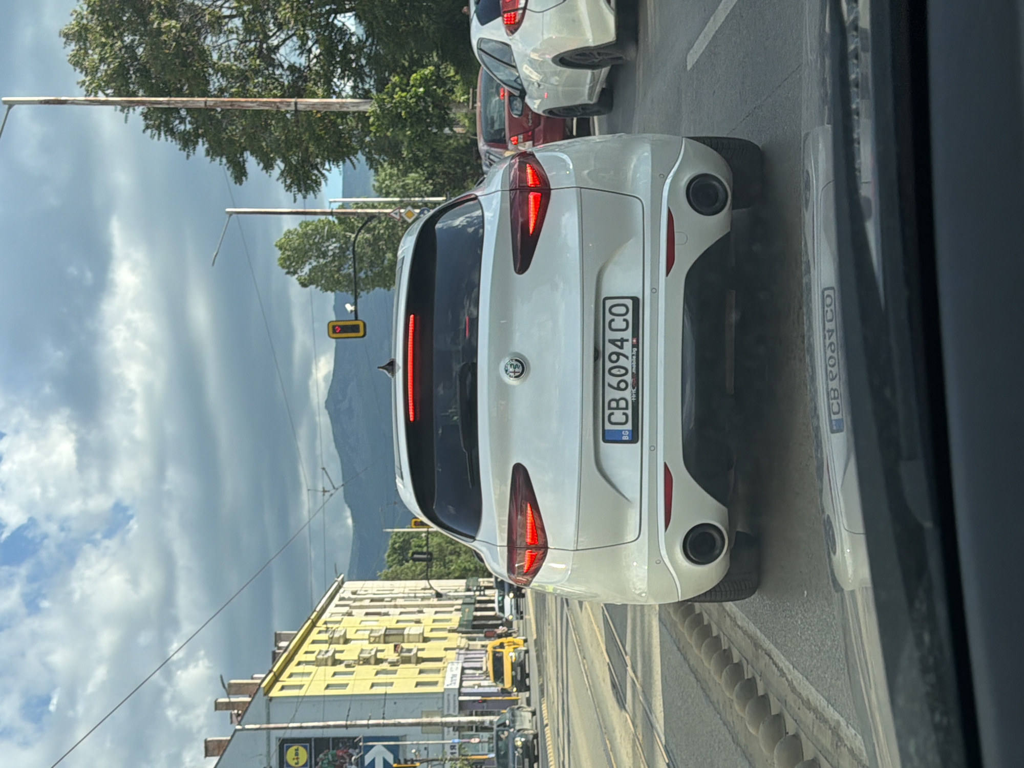 The image depicts the rear of a white Alfa Romeo Stelvio car with license plate "CB 6094 CO," which is a Bulgarian license plate. The car is located in Sofia, Bulgaria, as evidenced by the license plate prefix "CB". The image appears to be taken from within another vehicle, possibly a car. A traffic light can be seen in the background. The buildings in the background appear to be located near a LIDL store. In the background of the city is a mountain range. The sky is overcast. Other vehicles are visible in front of the Stelvio.