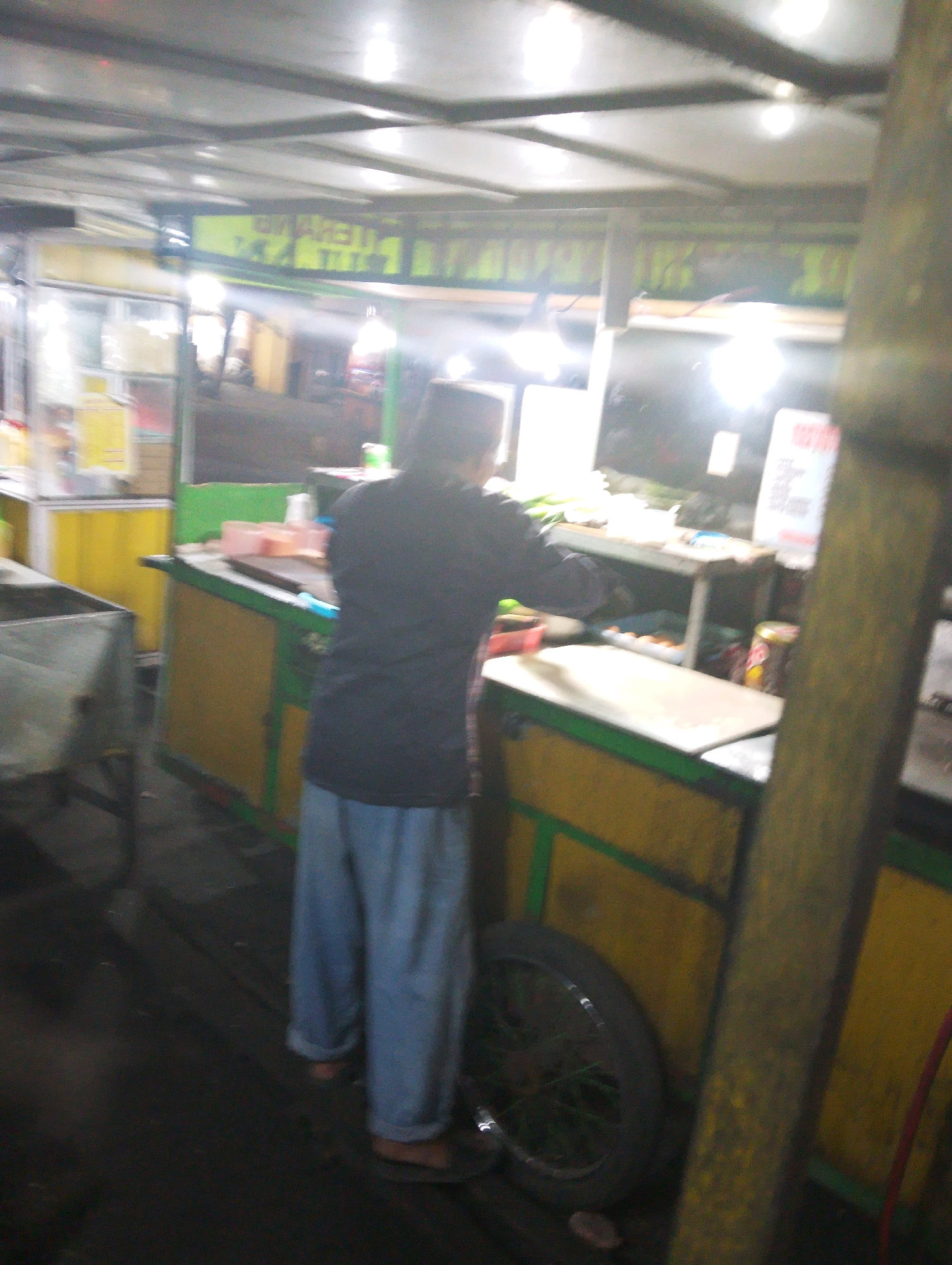 The image captures a dimly lit outdoor scene, likely a food stall or market stall, in Bangil, Indonesia. A person, viewed from behind, is wearing a dark long-sleeved shirt and light blue trousers. They are standing behind a food counter. The counter itself appears to be made of wood with a yellow and green color scheme. A prominent feature of the stall is a bicycle wheel, positioned on the ground to the right of the person and counter. Behind the counter, various items are visible, including what appears to be a whiteboard with writing, plastic containers, and possibly food items. The environment is enclosed by a roof structure, and the lighting suggests it may be evening or night. The overall impression is that of a functional, busy, but somewhat disorganized commercial space.