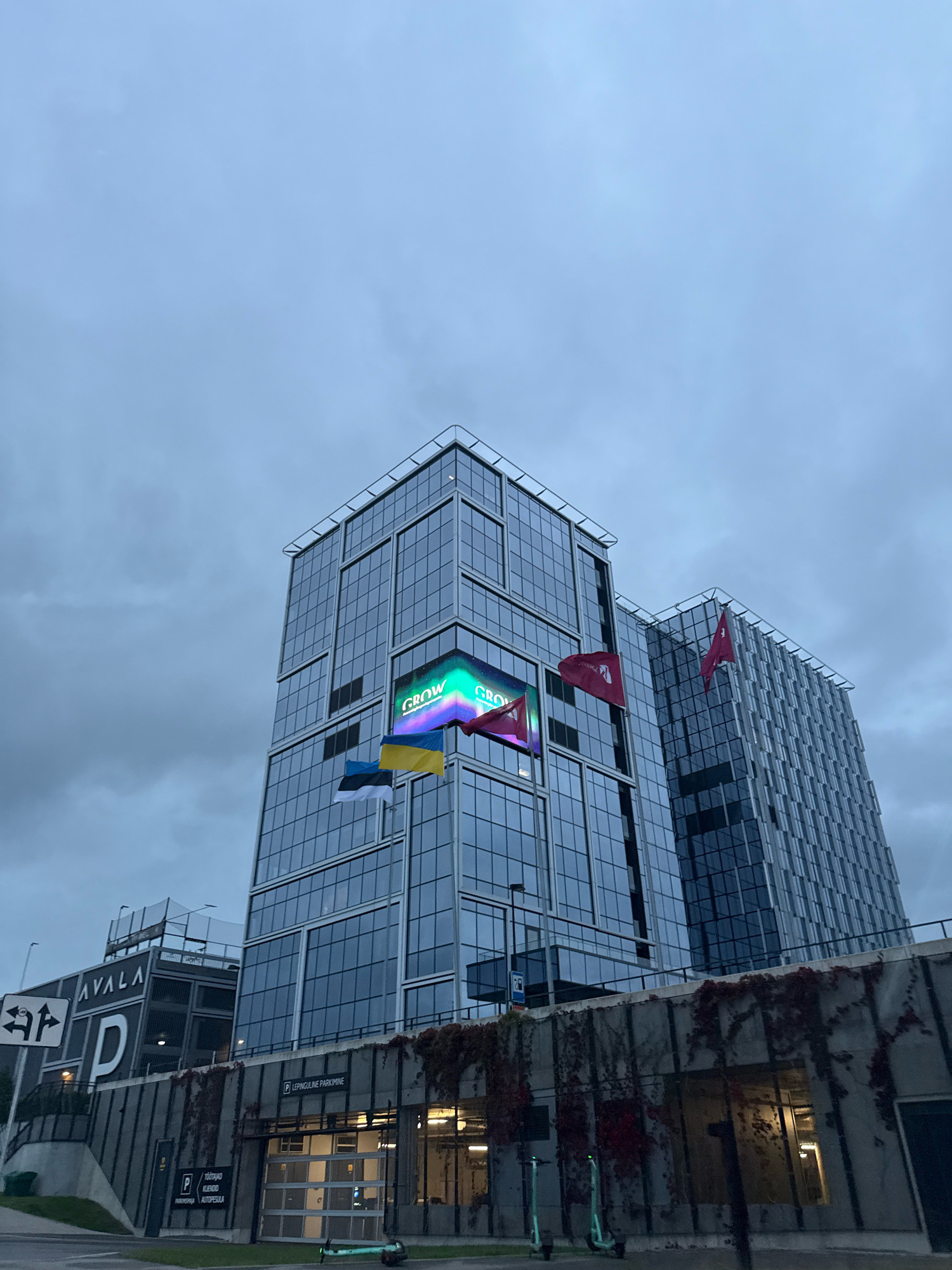 The image shows a modern glass-fronted office building in Tallinn, Estonia, under a cloudy sky. The building features an LED screen displaying the word "GROW" in colorful, illuminated letters. Several flags are visible: the Ukrainian flag, the Estonian flag, and two red flags with a white logo, likely representing a company. In the foreground, there is a concrete structure with an underground parking entrance, bearing the sign "P LEPINGULINE PARKIMINE" (Lease Parking). To the left, a multi-story parking garage is labeled "AVALA" with a large "P" symbol. Two electric scooters are parked near the parking entrance. The scene appears to be an urban commercial or business district.