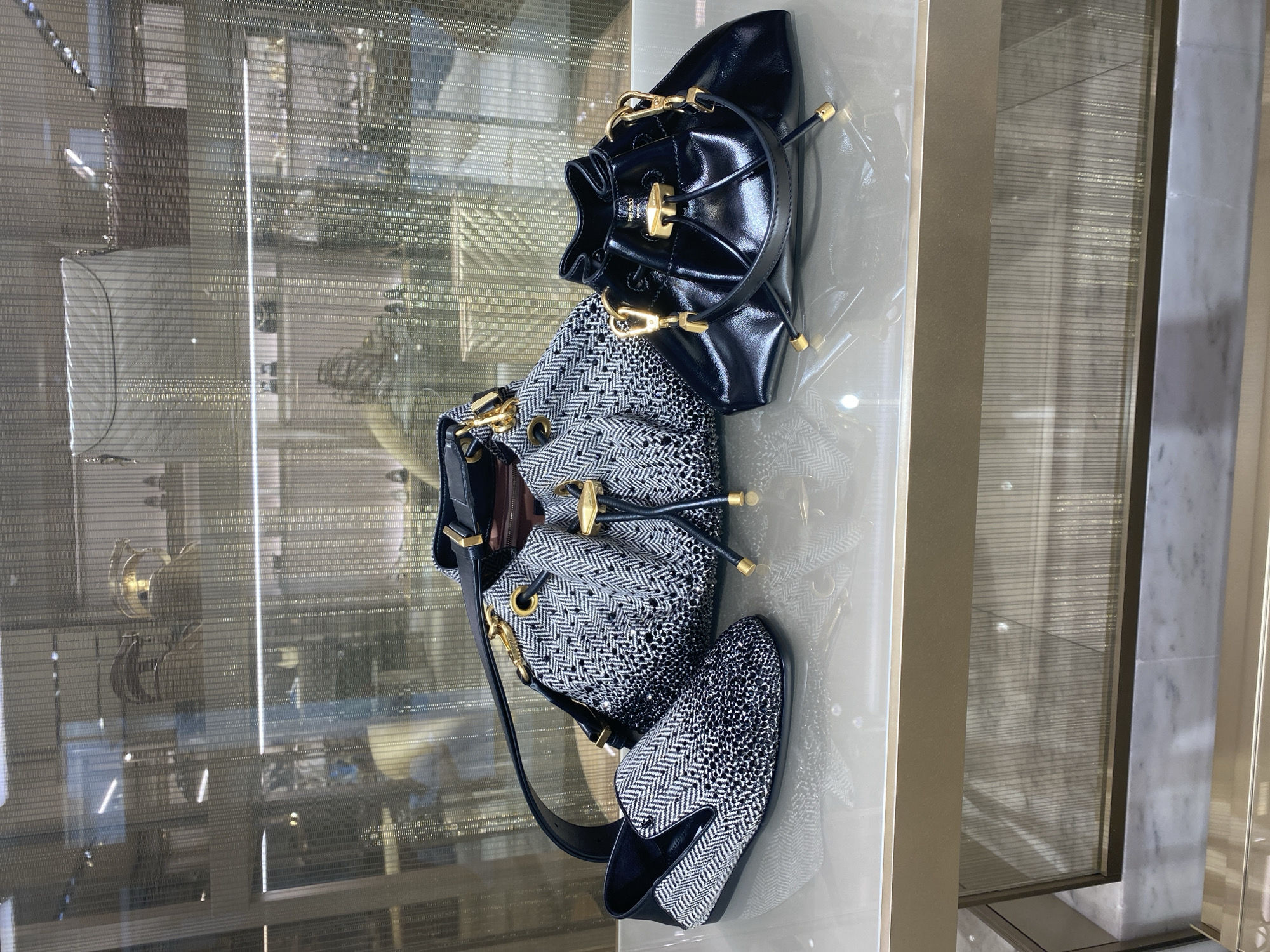 A display case in a retail environment showcases luxury accessories. In the foreground, on a reflective surface, sit two handbags and a pair of loafers. The leftmost item is a black and white patterned loafer with a pointed toe and a slight heel. To its right is a larger bucket-style handbag, also in a black and white zig-zag pattern, with a black leather strap and gold hardware. The third item, positioned to the right of the larger handbag, is a small, glossy black drawstring bag with a top handle and gold accents. The background of the display case is blurred, revealing the reflections of lights and other merchandise behind a textured screen. The overall scene is clean and well-lit, suggesting a high-end boutique setting.