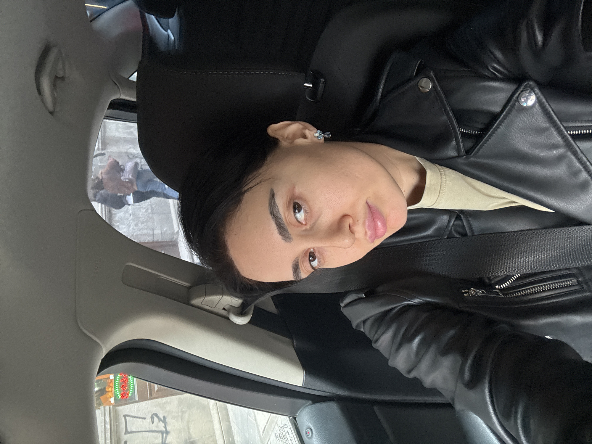 The image depicts a female subject seated in the front passenger seat of a vehicle. She has dark hair, brown eyes, and is wearing a black faux leather jacket over a light beige t-shirt, secured by a black seatbelt. A blue and silver floral-designed earring is visible on her right ear. The vehicle's interior features black upholstery and a light gray headliner and A-pillar, which displays an "AIRBAG" label.
Through the front driver's side window, a street scene in Tbilisi, Georgia, is visible. It shows a weathered building facade with dark graffiti and a blurred neon sign featuring red and green elements, indicative of a fruit or produce stand with oranges visible above. Through the rear passenger window, two blurred individuals are visible walking on a sidewalk. One person wears a dark jacket and light pants, while the other is in a reddish-brown top and blue jeans, carrying a bag. The setting is an urban street environment.