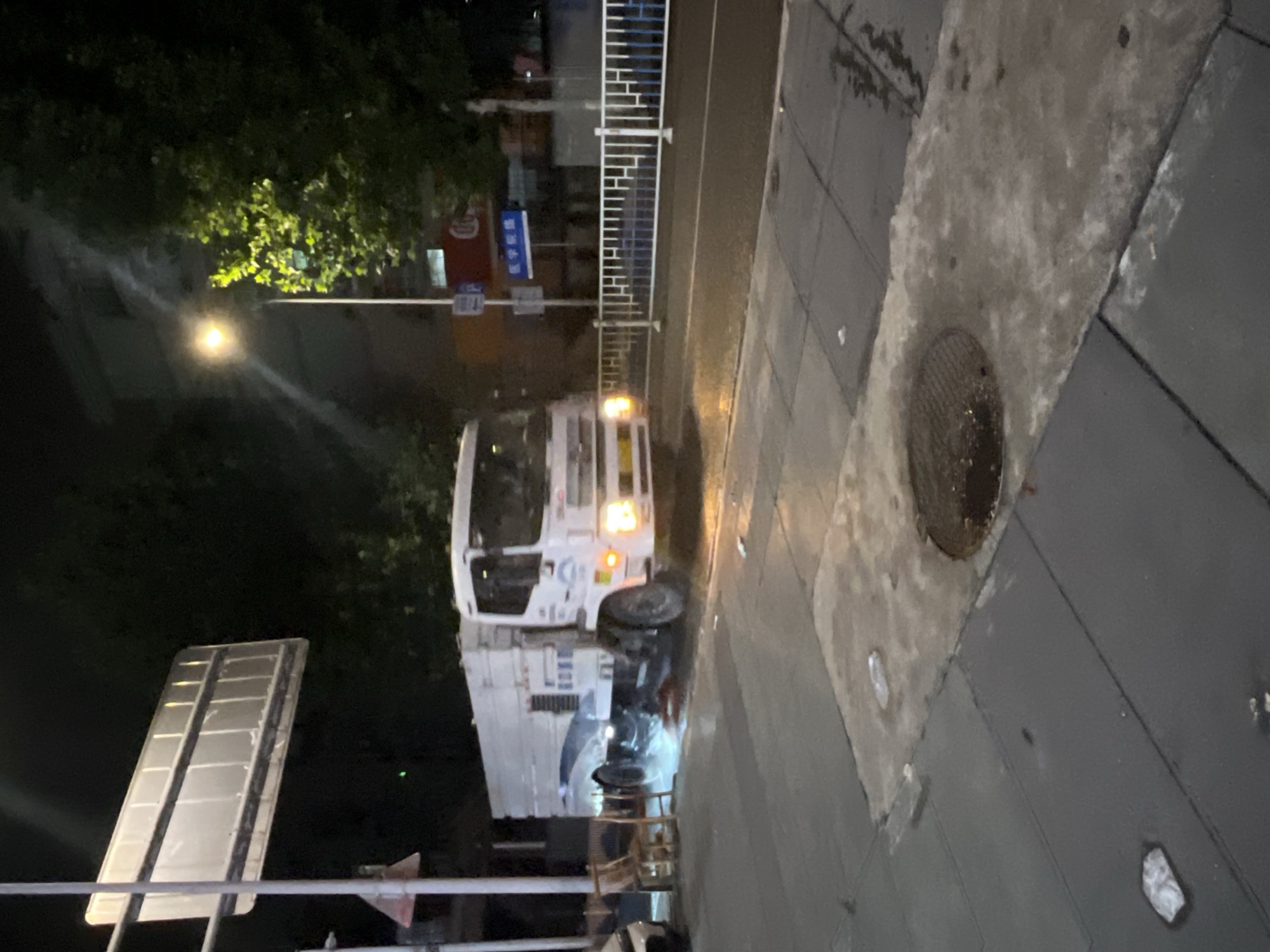 A nighttime scene in an urban area of Yuzhong District, China, depicts a wet street and sidewalk. A white utility truck, identified by markings including "B066" on its side, occupies the center of the road, angled slightly towards the right and displaying illuminated headlights. The truck is actively spraying or discharging a liquid from its left side onto the road surface, creating a distinct splash pattern.
On the left side of the frame, positioned on the dark gray paved sidewalk, are two wooden chairs and the partially visible front end of a white vehicle. The sidewalk also features a prominent circular manhole cover in the foreground and scattered pieces of minor litter.
Artificial lighting from streetlights illuminates the scene, casting reflections on the wet surfaces. A street light pole on the right side highlights a tree with green foliage and partially obscured building structures in the background. Street furniture includes a tall white pole with the rear of a large rectangular sign, a blue parking sign with a white "P," and a blue road sign bearing Chinese characters for "新宇宾馆" (Xinyu Hotel). Another sign in the distance displays "医学影像" (Medical Imaging). A white and blue pedestrian barrier lines the edge of the road on the right. No individuals are present in the image.
