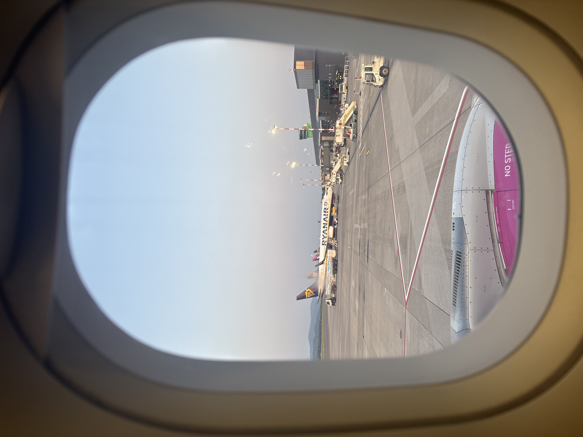 The image shows an airport tarmac scene as viewed from an airplane window. In the foreground, the engine of the airplane the photo is taken from is visible. Beyond that, multiple Ryanair airplanes are parked on the tarmac. An airport terminal building with a control tower is visible in the background, along with various airport service vehicles and mobile stairs. The sky is clear and blue, suggesting daytime. The scene is likely located at Orio al Serio International Airport (BGY) in Orio al Serio, Italy, as indicated by the presence of Ryanair planes, a major airline operating at that airport.