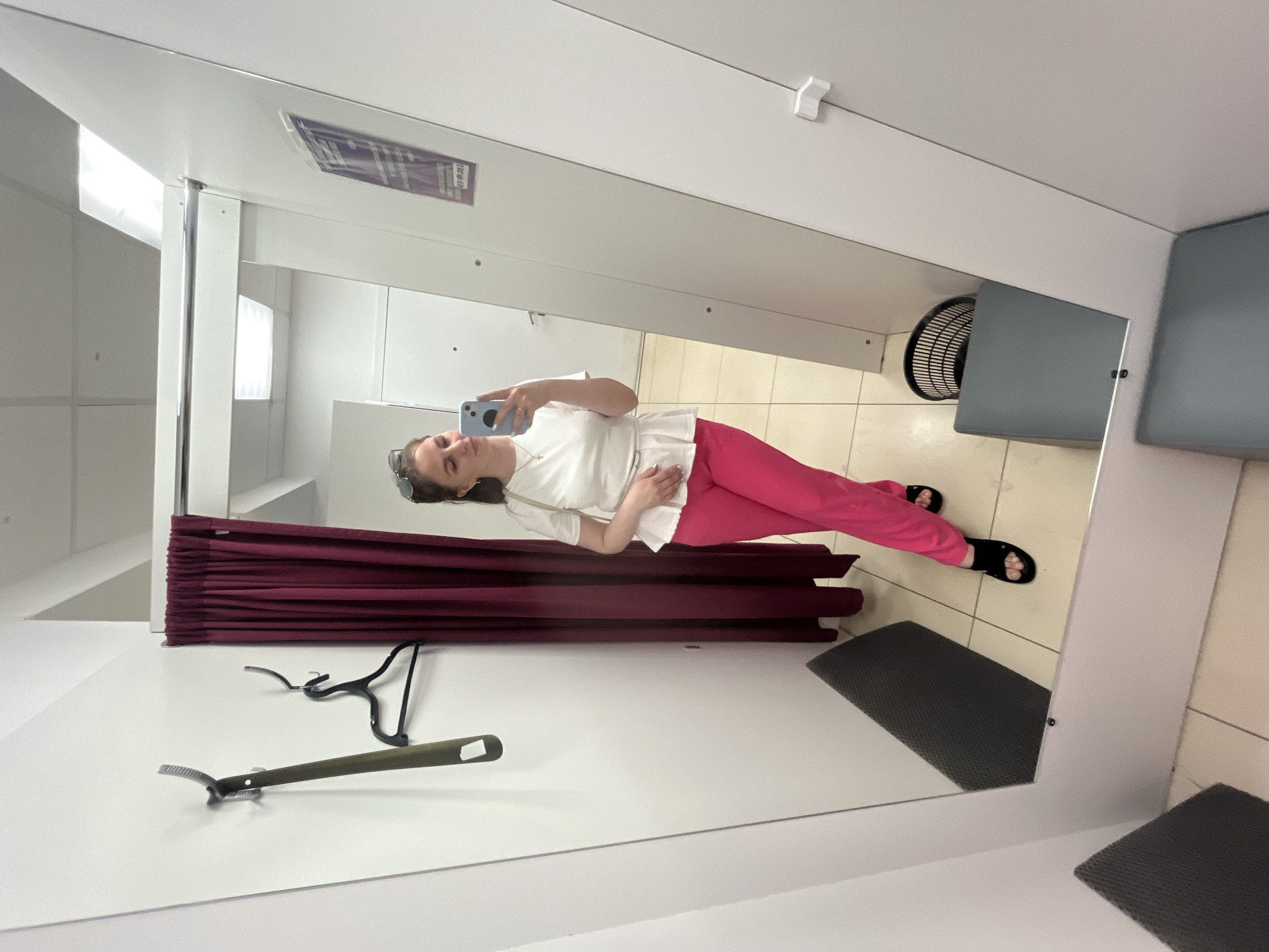 A woman is standing in a dressing room taking a selfie in a mirror. She is wearing a white top and pink pants and black sandals. She is holding a blue smartphone. The dressing room contains a burgundy curtain, a hanger, and a grey mat on a tiled floor. The mirror reflects the interior of the dressing room and the woman. There is a fan visible in the corner of the dressing room. There is no information in the image to verify the location within Izhevsk, Russia.