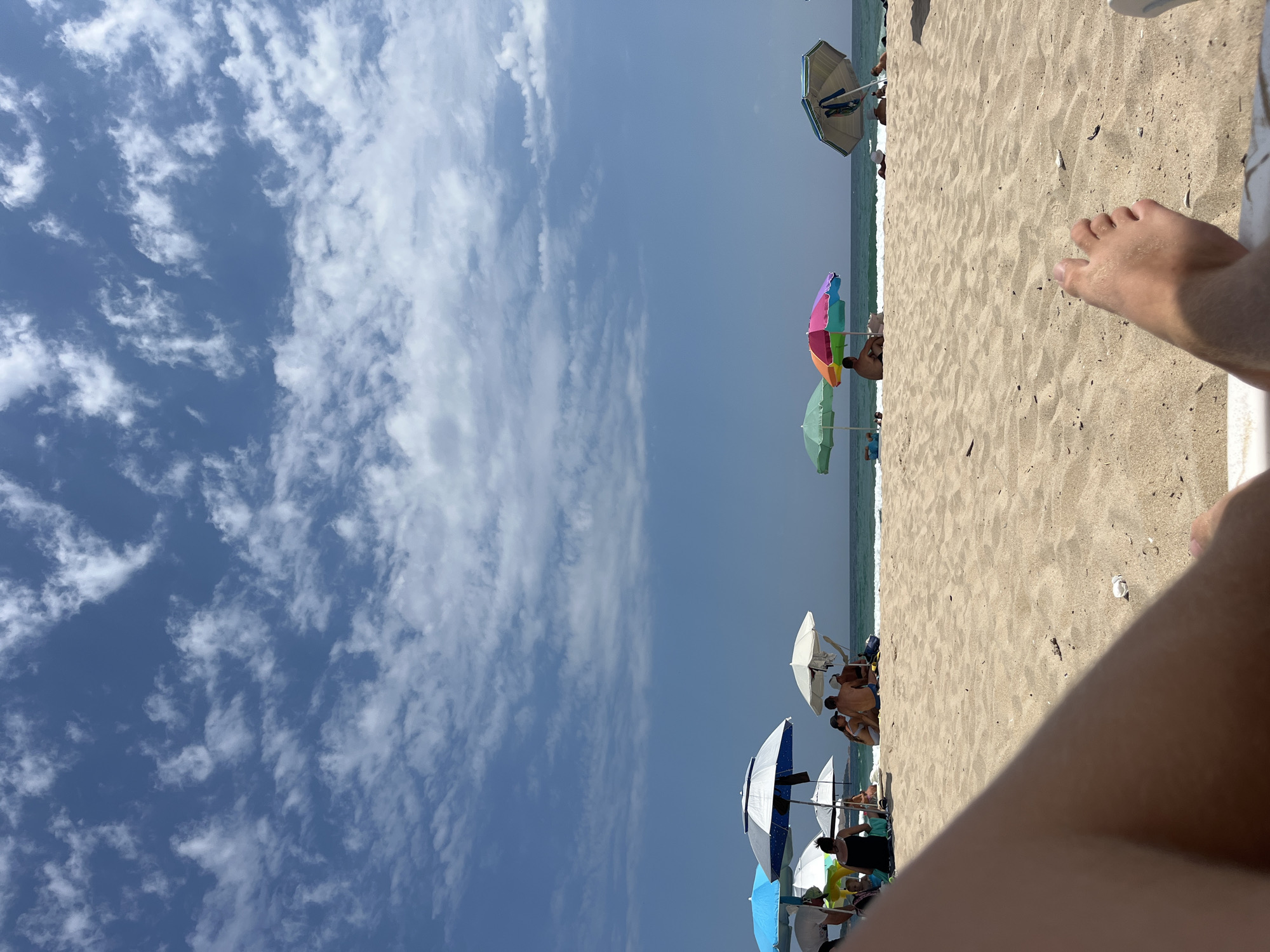 The image depicts a sunny day at a crowded beach, likely located in Primorsko, Bulgaria. The foreground is dominated by the sand of the beach and a person's leg and foot resting in what appears to be a lounge chair. In the midground, a line of beach umbrellas provides shade for beachgoers. Several umbrellas are visible, including a blue and white striped one, a white one, a rainbow-colored one, and a green one. People are seated or lying on the sand beneath these umbrellas, enjoying the seaside. Some individuals are partially visible, suggesting a relaxed atmosphere. The background features the calm sea meeting the horizon under a bright blue sky scattered with white, wispy clouds. The overall scene conveys a typical summer day at a popular beach resort.