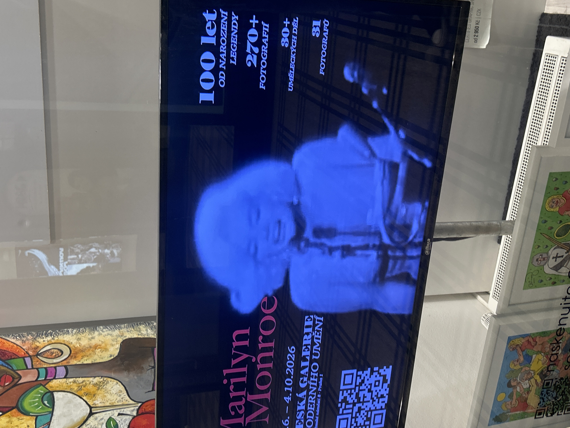 The image shows a digital display showcasing an exhibition about Marilyn Monroe. The display features a large, blue-tinted image of Marilyn Monroe herself, alongside text announcing "Marilyn Monroe" and exhibition details. The text is in Czech, reading "100 let OD NAROZENÍ LEGENDY" (100 years since the birth of the legend), "270+ FOTOGRAFIÍ" (270+ photographs), "30+ UMĚLECKÝCH DĚL" (30+ works of art), and "31 FOTOGRAFŮ" (31 photographers). Below the name Marilyn Monroe, there is exhibition information that includes dates ".6. - 4.10.2026" and the name of the venue: "ČESKÁ GALERIE MODERNÍHO UMĚNÍ" (Czech Gallery of Modern Art). A QR code is also visible on the lower left of the display. The background of the display has a subtle checkered pattern. The digital display is mounted on a stand, and the bottom of the image shows the top of two framed artworks below the display, one depicting a group of people in a beach-like setting and another showing a knight. In the lower right corner of the display screen, there is a price listed as "od 2 950 Kč | CZK", indicating the price in Czech Koruna. The exhibition appears to be taking place in Prague, Czechia, given the Czech language used on the display and the mention of "Praha 1" (Prague 1) within the gallery's address.