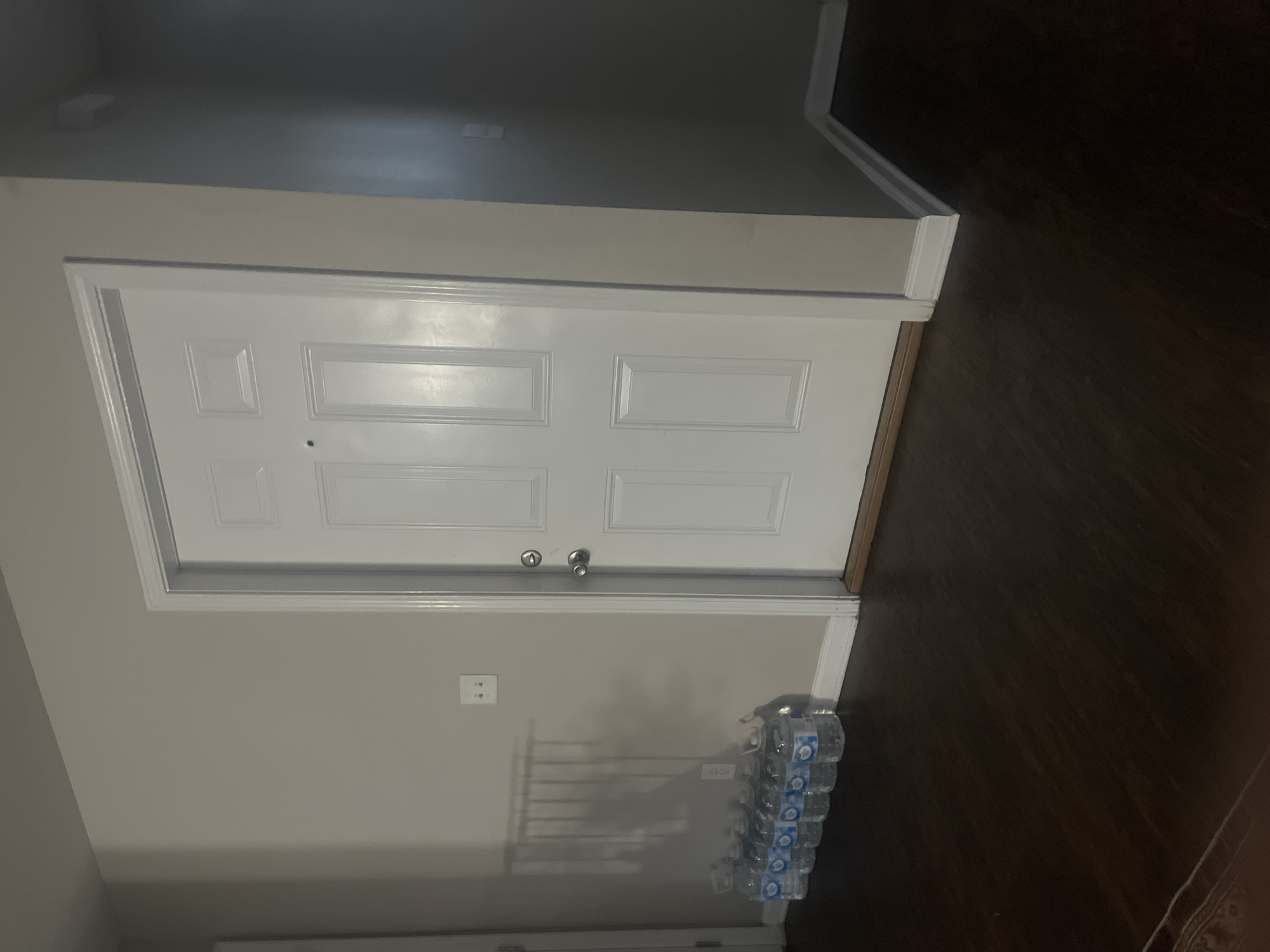 A white paneled interior door is positioned in the center of the frame. To the left of the door, a row of clear plastic water bottles with blue labels are arranged on the dark wooden floor. A light-colored wall with a visible light switch is to the left of the door. The floor is a dark brown wood laminate. The scene appears to be indoors, likely in a residential setting.