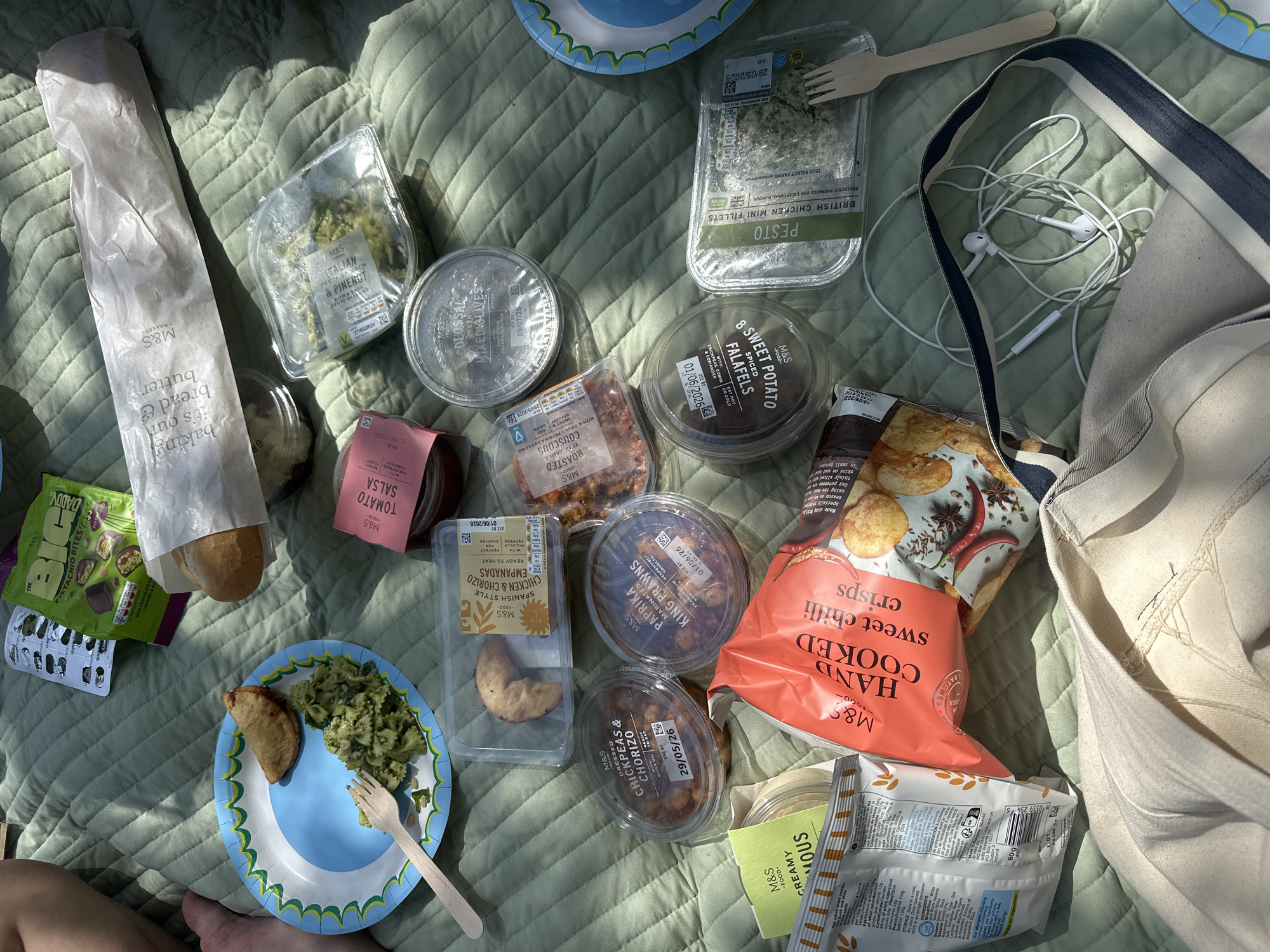 The image shows a picnic spread laid out on a patterned green blanket, likely outdoors. A variety of M&S food items dominate the scene, including: a bread bag, pistachio bites, tomato salsa, vegetable couscous, chicken and chorizo empanadas, chickpea and chorizo dish, paprika marinated king prawns, sweet potato falafels, pesto British chicken mini fillets, pasta spinach and pinenut salad, sweet chilli crisps and creamy hummus. There's also a container of mixed olives. A blue and white patterned plate is visible, holding pasta salad and an empanada, with a wooden fork. A pair of white earphones are present near a partially visible bag. Some items have "use by" dates in late May and early June 2026. Part of a person's leg is visible in the lower left corner. The items suggest the picnic took place in the United Kingdom, based on the M&S branding. The location is likely a park or outdoor area, but the specific location in London is not discernible from the image.