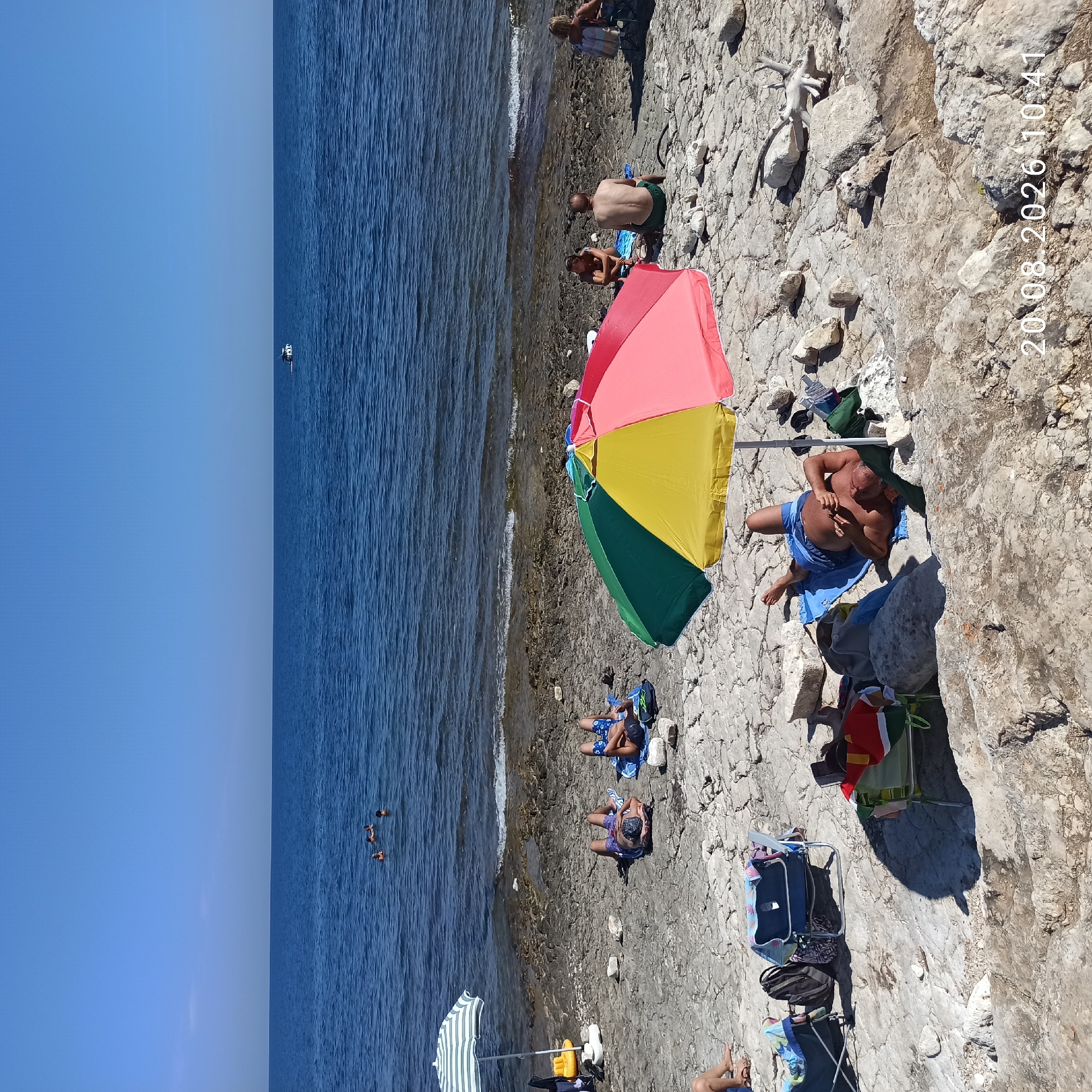 The image depicts a sunny day at a rocky beach near Molfetta, Italy. Several people are scattered across the shore, some sunbathing on towels, others sitting in chairs or in the shallow water. Two individuals are visible in the sea, along with a distant boat. A colorful umbrella provides shade for two people who are relaxing on the rocky ground. Beach chairs, bags, and other personal items are also present. The water is calm and blue, meeting a rocky coastline with sparse vegetation. The sky is clear and blue, indicating fair weather. A watermark of "20.08.2026 10:41" is present in the bottom right corner of the image.