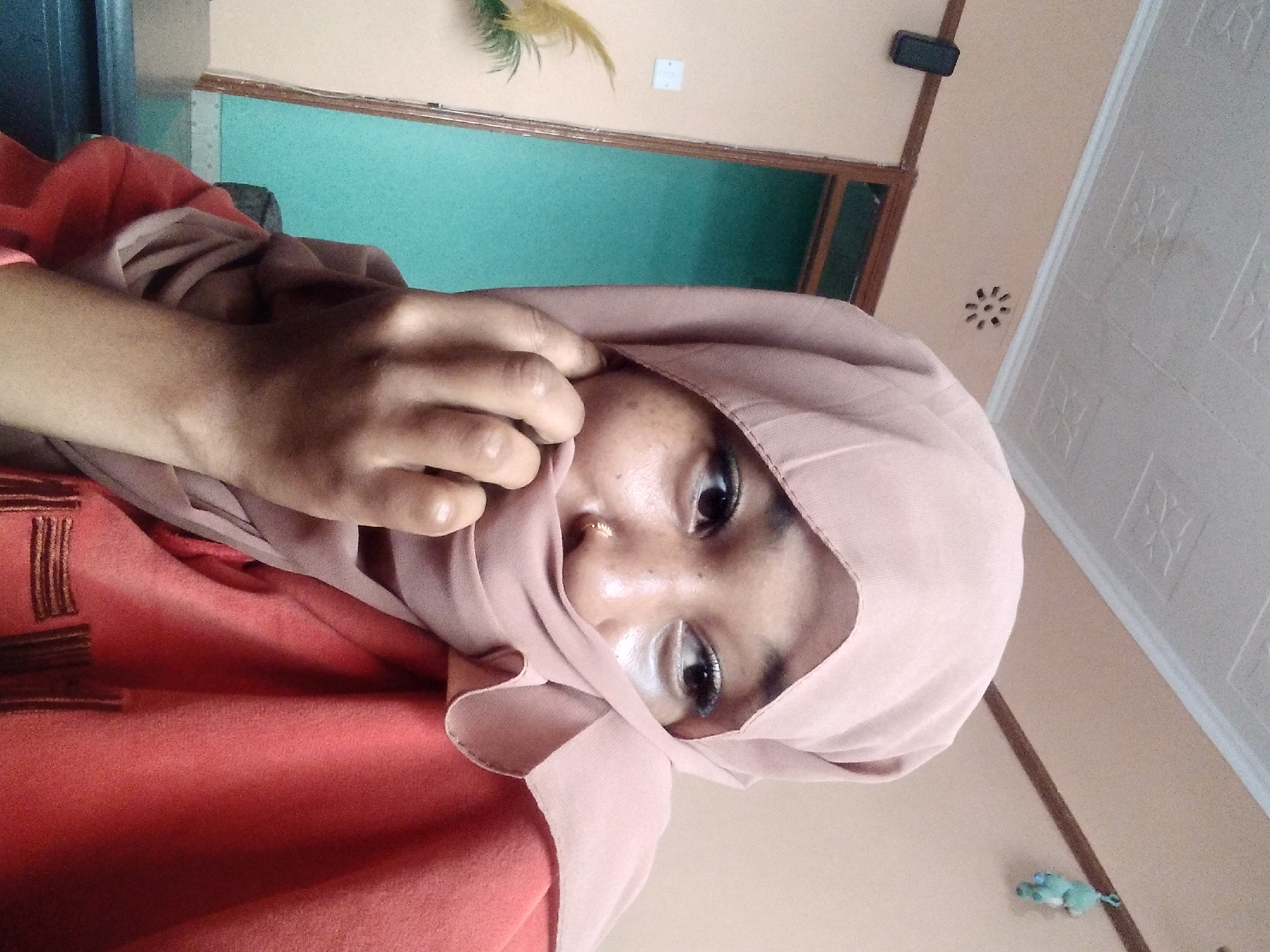 The image depicts a close-up of a young woman wearing a light pink hijab and an orange top. She is partially covering her face with the hijab and her left hand is near her mouth. Her eyes are visible, and she appears to be looking to her right. A nose ring is visible. The background suggests an indoor setting with a light-colored wall, a door frame, and a ceiling with textured tiles. A small decorative item, possibly a toy, is hanging on the wall in the upper right corner. The overall lighting is soft, and the image has a slightly grainy quality. The location is identified as the city of Nyeri, Kenya.