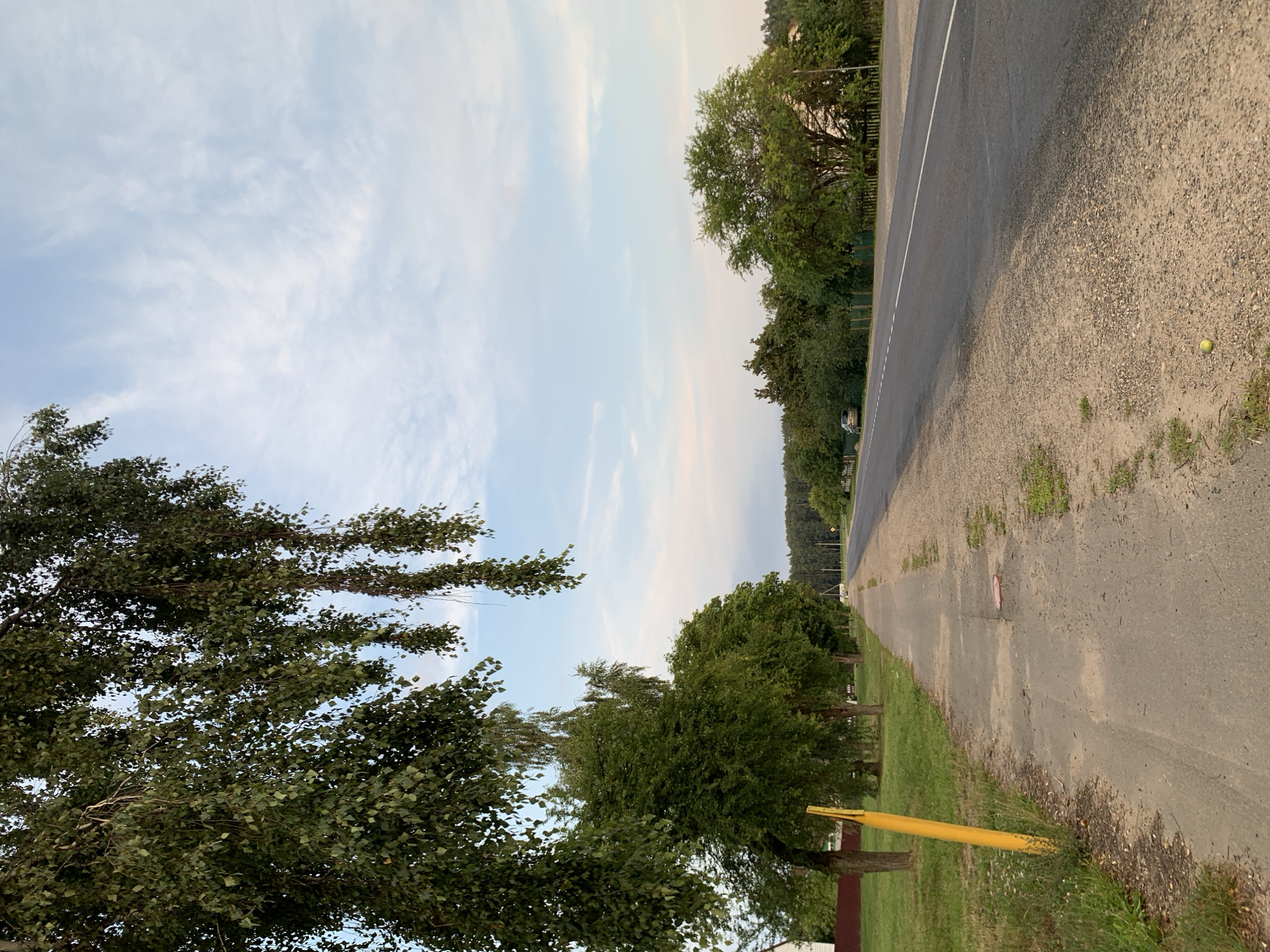 The image depicts a rural road scene in Kapyĺ, Belarus, under a cloudy sky. The foreground shows a paved sidewalk with a bright yellow pole on the left. Beyond the sidewalk, a row of green trees lines the road. The road itself is asphalt with a white line marking the center. In the distance, more trees and vegetation are visible, forming a dense forest. A car is parked on the right side of the road. The overall impression is a quiet, somewhat secluded, rural landscape.