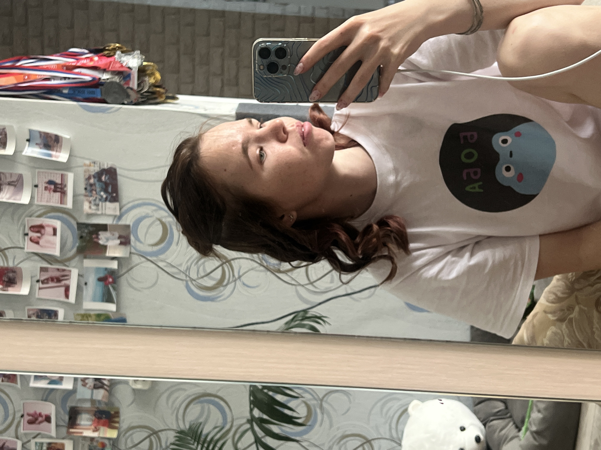 A young woman takes a selfie in a mirror. She has medium-length brown hair with red highlights and is wearing a white t-shirt with a blue frog graphic on it. The woman is holding a smartphone with a textured blue case in her left hand. She is looking at the phone, presumably to frame the shot. A white earbud wire is visible near her shoulder. In the background, a wall with a wallpaper featuring blue circular patterns and a collection of small photographs is visible. A shelf above the photos holds a pile of medals. Below the mirror, a glimpse of greenery, possibly a plant, and a small white bear figurine are seen. The location is Chaikovsky, Russia, based on the objects in the background.